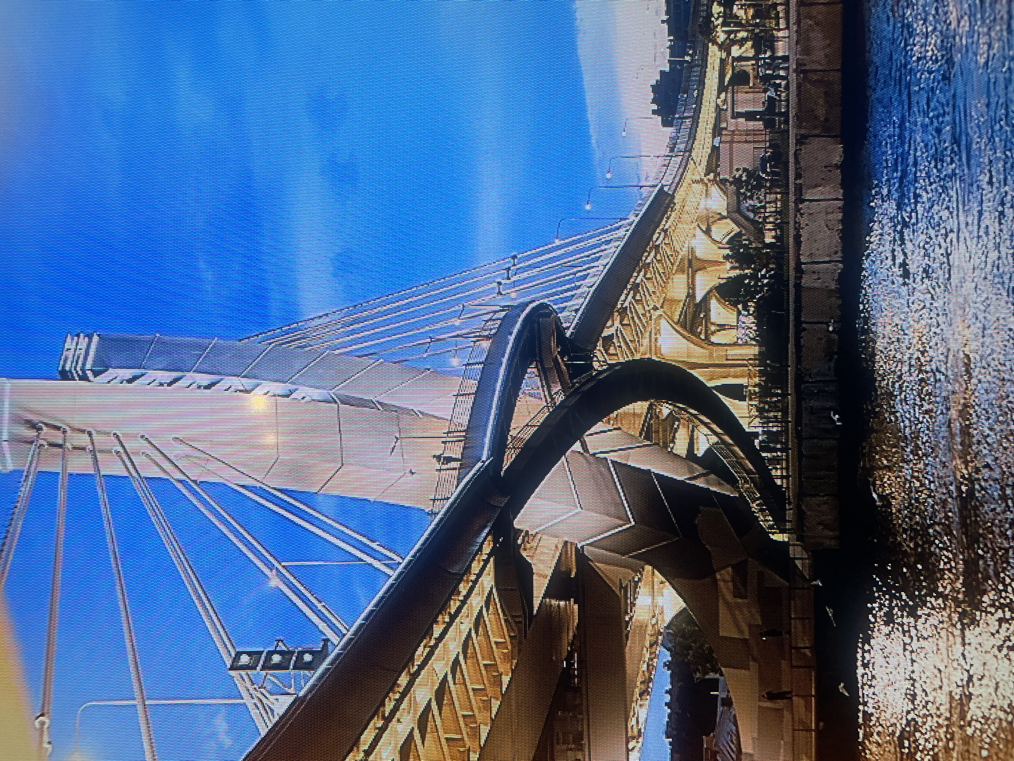 The image shows a cable-stayed bridge in the city of Mytishchi, Russia. The bridge has a distinctive design with a tall central pylon and numerous cables supporting the roadway. The roadway itself appears to be illuminated, casting a warm glow on the structure. Below the bridge, there is a body of water, possibly a river or canal, with reflections of the bridge and the sky on its surface. The sky is a clear blue with some wispy clouds. On the pedestrian walkway of the bridge, there are a few silhouetted figures. The overall scene suggests a modern urban landscape with a focus on impressive architectural engineering.