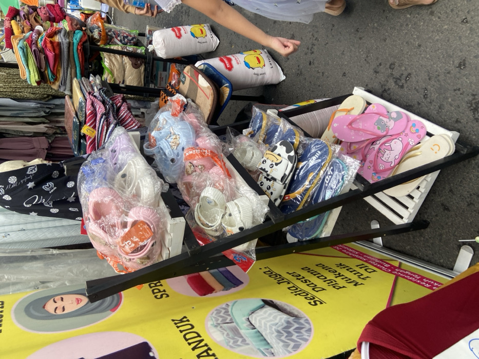 The image shows a street vendor selling flip flops and slippers in Kecamatan Sidoarjo, Indonesia (latitude: -7.442, longitude: 112.705). The vendor has a four-tiered rack stacked with pairs of assorted footwear. There are two people in the scene, a female customer and a street vendor (unseen). The customer is wearing a pink and white dress and is holding a yellow and blue shopping bag. The vendor's stall is set up on a paved sidewalk. The rack has multiple shelves with various sizes, styles, and colors of flip flops and slippers. Some of the footwear features cartoon characters printed on them. The environment has a clutter of various objects, including rolled-up fabric items, a yellow and blue plastic bag, a colorful mat, a yellow-and-blue plastic bag with a duck logo, and other items that are not visible in the image. The vendor is not visible in the picture.