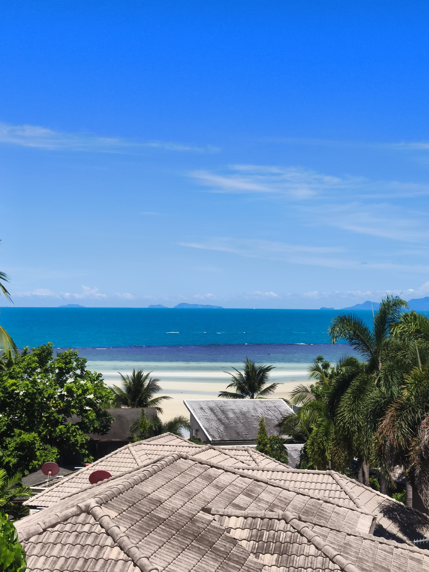 This is an elevated, eye-level, medium shot of a tropical beach scene. The foreground is dominated by the tiled roofs of several buildings, with two red satellite dishes visible on one roof. Lush green foliage and palm trees are scattered throughout the middle ground, partially obscuring other buildings and more tiled roofs. The background features a bright, sandy beach that slopes into a calm, turquoise ocean. In the distance, a hazy horizon is punctuated by several mountainous islands. The sky is a vibrant, cloudless blue. The scene suggests a warm, sunny day in a coastal resort area, likely in Baan Thong Po, Thailand, given the tropical vegetation and architecture. There are no people or animals visible in the image.