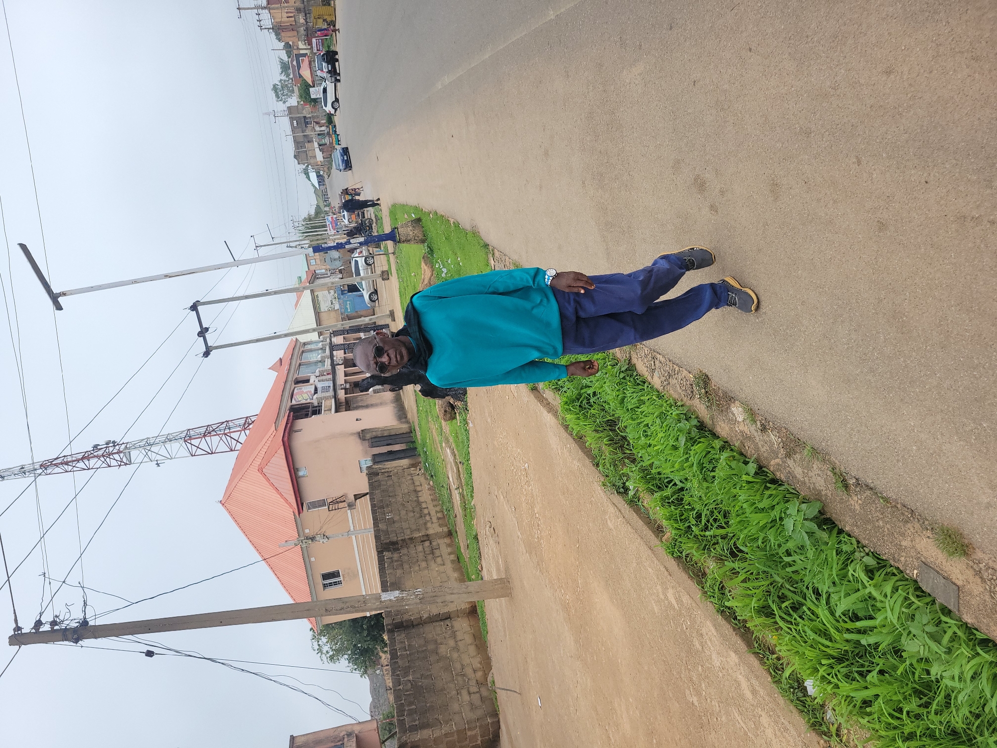 The image captures a street scene in Jos, Nigeria, on an overcast day. A man, appearing to be in his 60s with a bald head and wearing sunglasses, stands on a paved sidewalk next to a grassy drainage ditch. He is dressed in a teal long-sleeved shirt, dark blue pants, and dark athletic shoes with orange accents. A dark scarf is draped around his neck. To the left of the man, a concrete wall separates the sidewalk from a dirt area with some vegetation. Beyond the wall, a building with a salmon-colored roof and cream-colored walls is visible, along with a tall communication tower. The street in front of the man is asphalt, with several cars and motorcycles parked or driving in the distance. The environment suggests an urban setting with utility poles and streetlights lining the road.