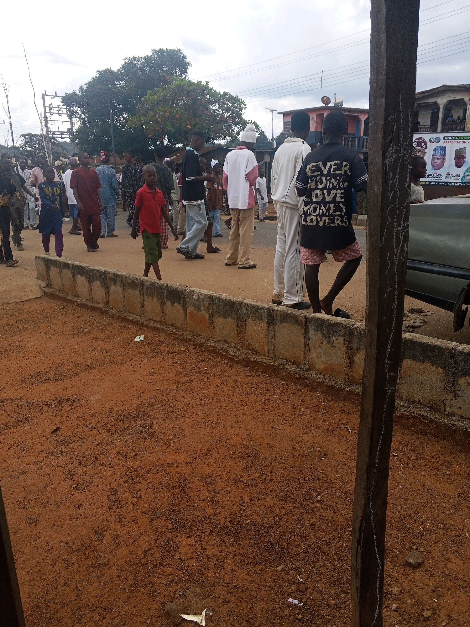 The image depicts a street scene in Egbedi, Nigeria. A group of people, including men, women, and children, are gathered and walking along a dirt road. The road is bordered by a low concrete barrier. Several individuals are dressed in traditional Nigerian attire, while others wear casual clothing. One boy in the foreground wears a red t-shirt and green shorts, and another young person is seen in a black t-shirt with white lettering that reads "NEVER HATING LOVE MONEY LOVERS". In the background, buildings are visible, including one with a banner displaying portraits of political figures, suggesting a possible campaign event. Trees and utility poles are also present, indicating an urban or semi-urban environment. The sky is overcast.