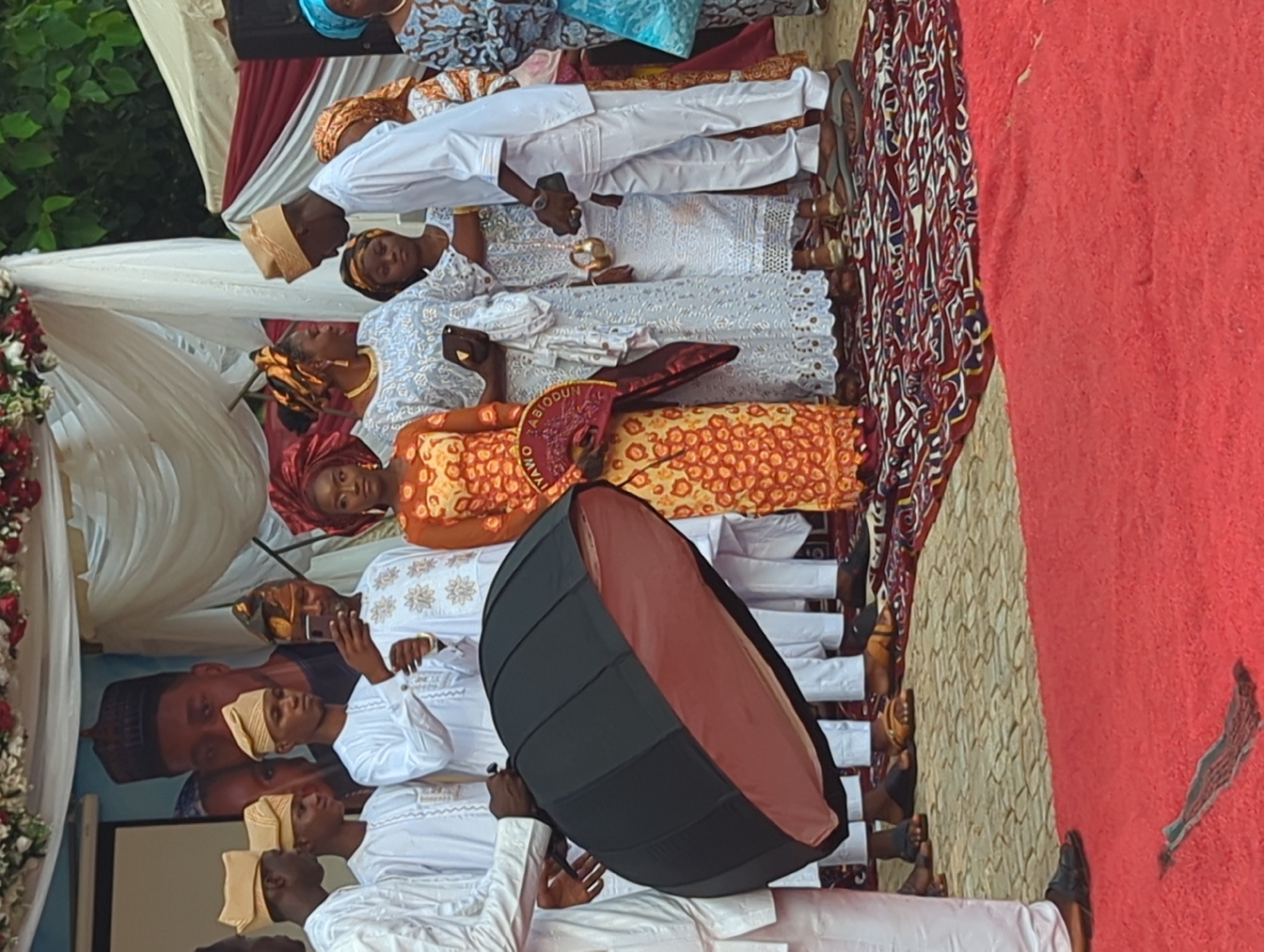 In Kabala, Nigeria, a group of individuals participates in what appears to be a formal event, likely a traditional wedding ceremony, indicated by the 'IYAWO ABIODUN' fan. The scene unfolds on a stage or raised platform covered with a patterned rug and a red carpet.
On the far left, a person in white traditional attire, visible from the torso down, holds a large black octagonal photography softbox with an orange interior, angled towards the central figures. To their right, four young men in white traditional garments and matching light-colored caps are present. One of these men, wearing a darker patterned cap, holds a smartphone up, seemingly capturing a photograph or video.
The central figure is a woman in a vibrant orange and yellow patterned dress and a matching headwrap (gele). She holds a red and gold fan inscribed with "IYAWO ABIODUN" (meaning "Wife Abiodun" in Yoruba). To her immediate right, another woman in a light blue and white lace dress with a gold headwrap holds a dark clutch bag and looks towards her right. A young girl in white lace and a patterned headwrap stands partially visible behind this woman.
Further to the right, a man in white traditional attire and a light-colored cap looks down at a small, ornate golden vessel held by a woman in white lace, whose face is mostly obscured. The man holds a dark smartphone in his left hand. Another woman in a blue and brown patterned dress and blue headwrap stands on the far right.
The background features white and red drapery, with red and white floral arrangements along the top. A large poster depicts a man in traditional Nigerian attire, with another person partially visible behind him. The ground beyond the red carpet appears to be paved with light-colored interlocking stones. The participants are wearing a mix of sandals and closed shoes.