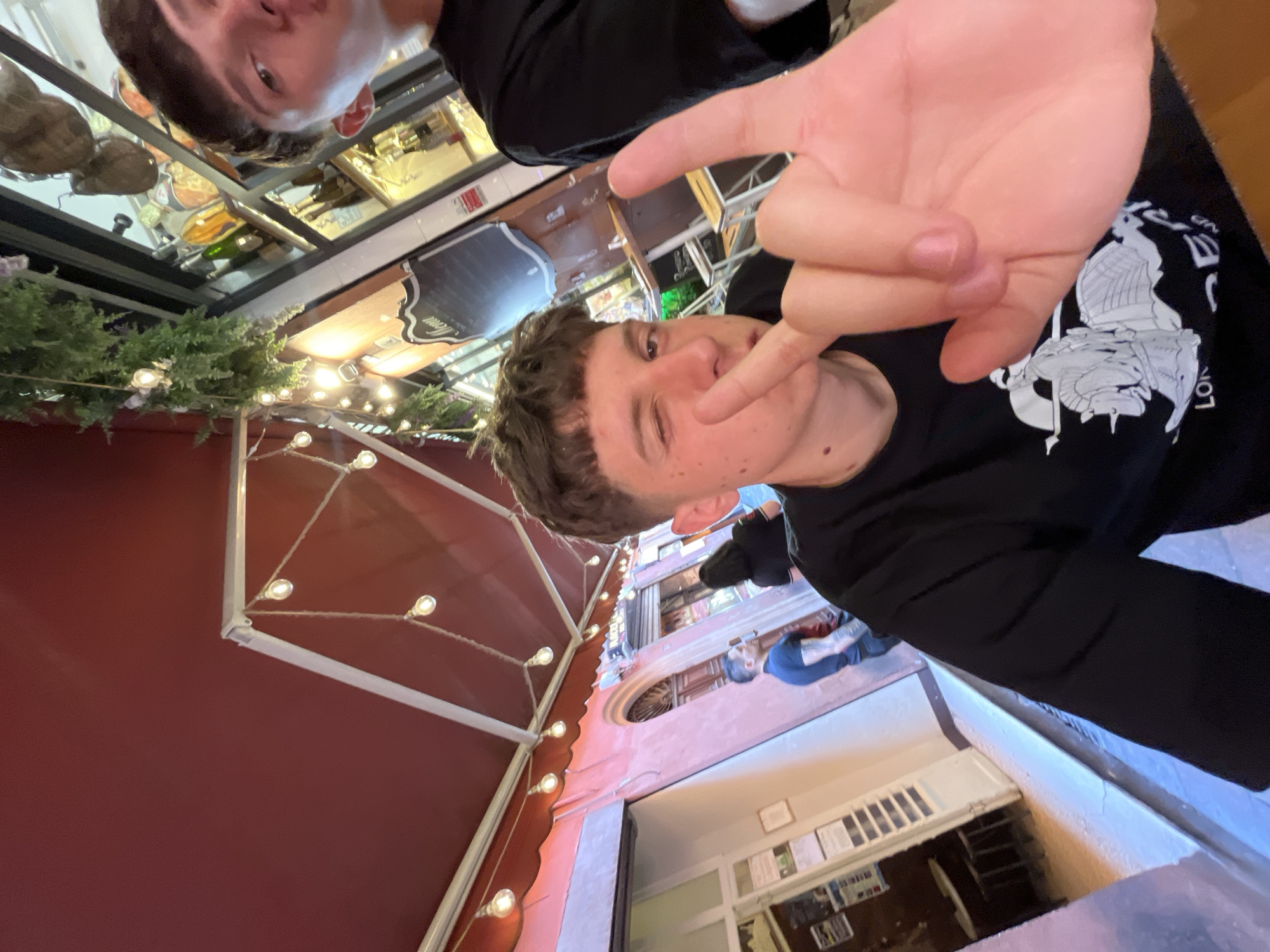 The image is taken in Bologna, Italy. It depicts a selfie taken by a young man with light brown hair, wearing a black long-sleeved shirt with a graphic design on it. He is winking and making a hand gesture with his right hand close to the camera. Another person is partially visible in the upper part of the frame. The scene is in an outdoor restaurant or cafe setting. Behind the subject, the architecture of Bologna is visible. A red awning adorned with lights is above, and beyond that, there is a view down a narrow street lined with buildings, with other people visible in the distance. The image is slightly rotated.