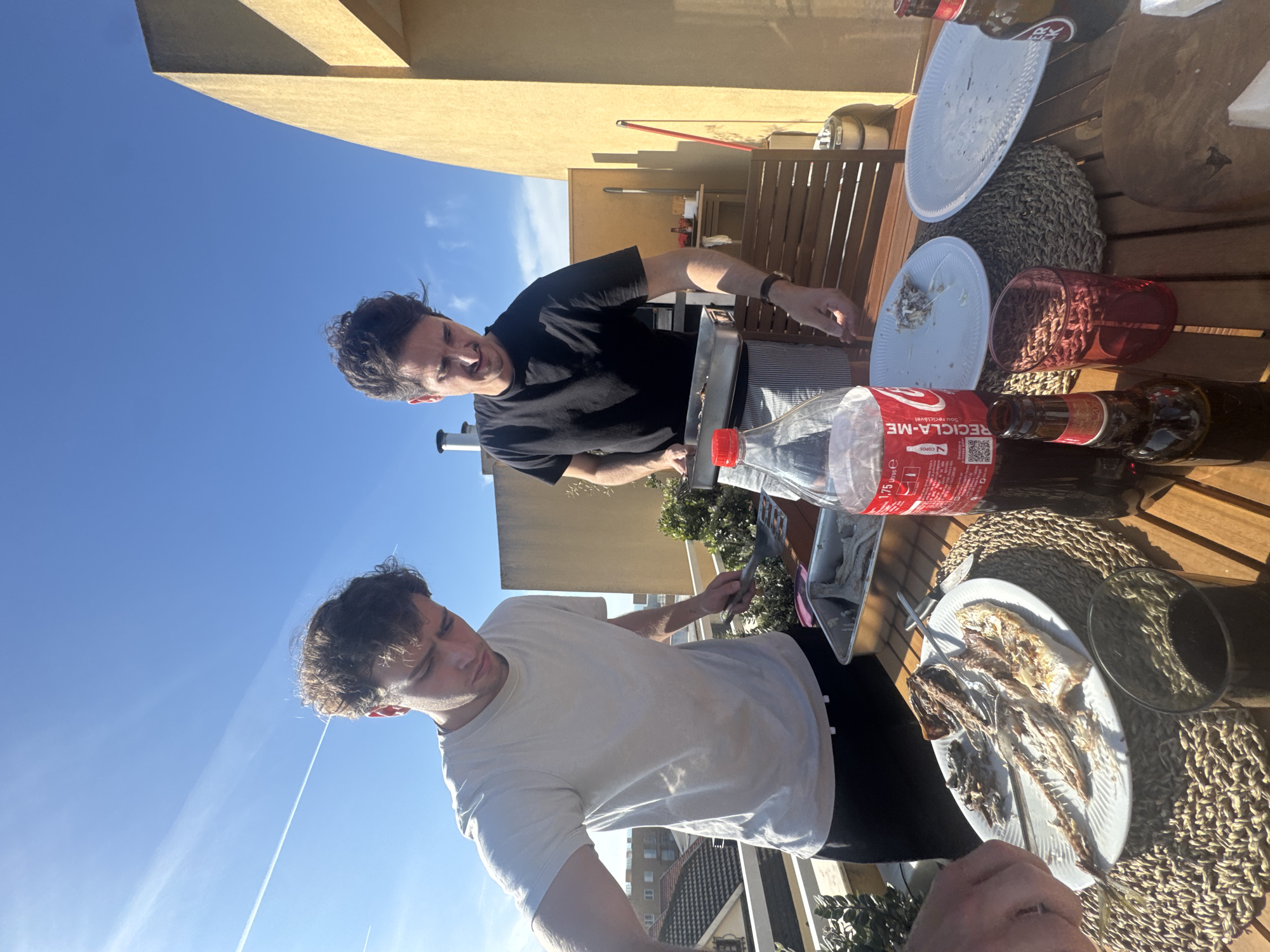 Two men are on a rooftop in Vila Nova de Gaia, Portugal, preparing a barbecue. One man, wearing a white t-shirt and black pants, is holding a plate with fish bones, having just eaten. The other man, wearing a black t-shirt, is tending to the barbecue grill. There is a wooden table with placemats, plates, glassware, and a bottle of Coca-Cola. In the background, the urban landscape of Vila Nova de Gaia is visible under a clear blue sky.