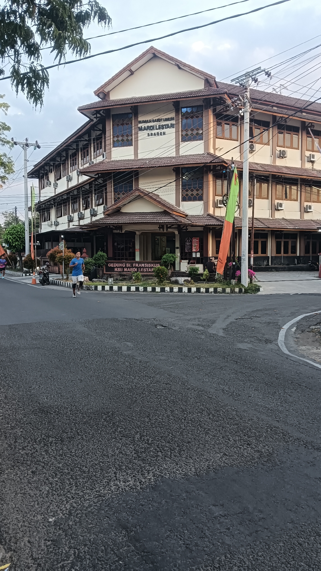 The image shows the RSU Mardi Lestari hospital in Sragen, Indonesia. A man is jogging on the road in front of the building. The hospital is a multi-story structure with a brown tiled roof and white walls. There are several windows on each floor, some with decorative patterns. A green and orange flag is hanging from a pole in front of the building. The road is paved and appears to be well-maintained. The sky is overcast.