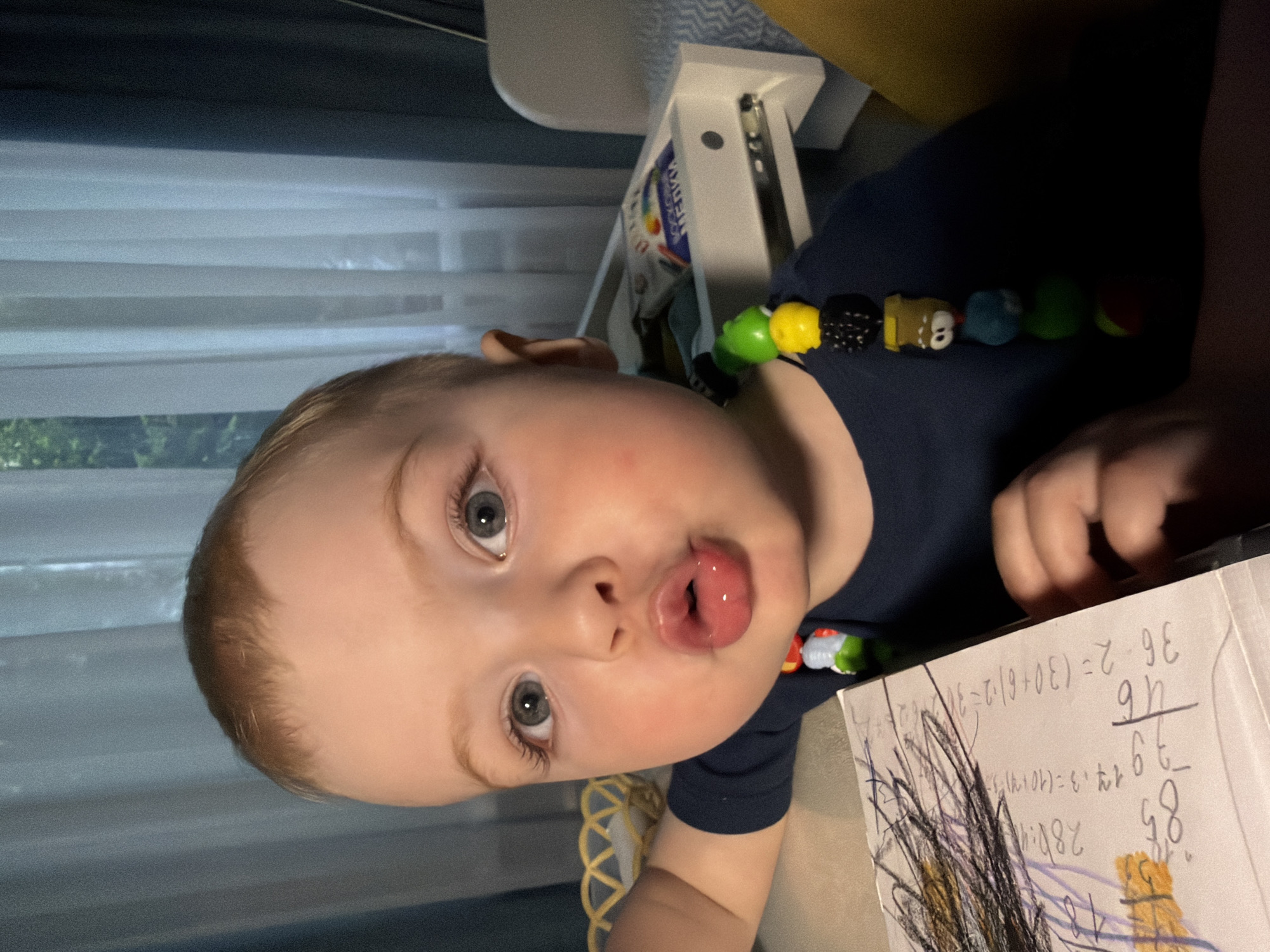 A close-up, vertical, outdoor shot shows a young Caucasian child with light brown hair and grey eyes posing with their mouth pursed. The child is wearing a dark blue t-shirt adorned with a colorful beaded necklace featuring cartoon characters. The child is leaning on a desk with a notebook open in front of them, showcasing what appears to be handwritten math problems and drawings. The background is a domestic indoor setting with white curtains and a white piece of furniture with drawers. The lighting suggests it's daytime. The child's pose and expression indicate a playful interaction with the camera. The image was taken in Votkinsk, Russia.