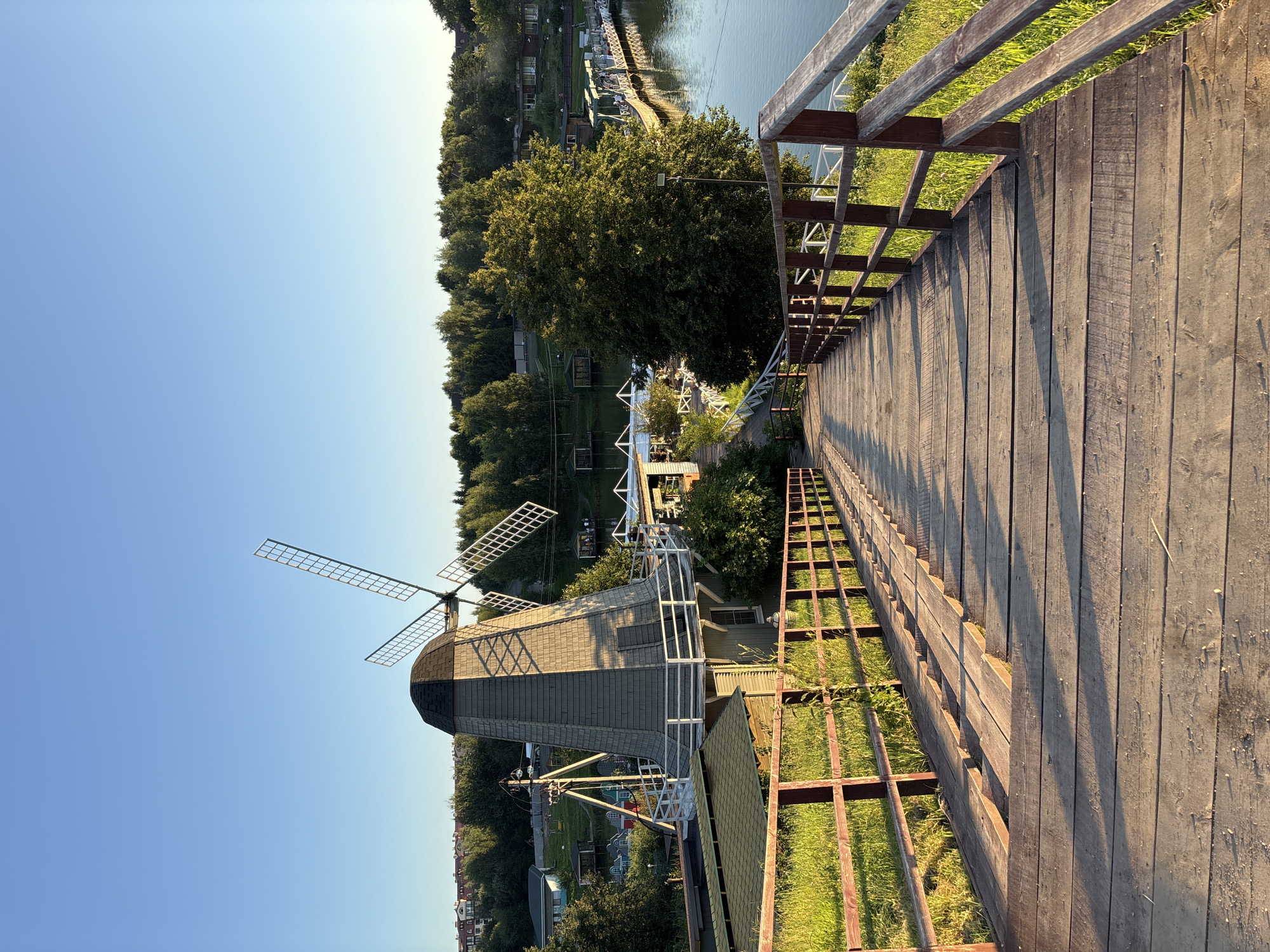The image depicts a wooden staircase leading down towards a body of water with a windmill in the background. The staircase has wooden railings on both sides, and the steps are weathered and uneven in places. To the left of the staircase, there is a grassy area with a small building and some trees. The windmill is a traditional design with a dark roof and light-colored body, and its sails are visible. Beyond the windmill, there are more trees and some buildings, suggesting a residential or recreational area. The water is calm and reflects the clear blue sky. The scene appears to be set in a rural or semi-rural location, likely a park or a lakeside resort. The image was taken from the top of the staircase, looking downwards. The lighting suggests it is late afternoon or early evening, with the sun casting long shadows across the staircase. The location is identified as Romashkovo, Russia.