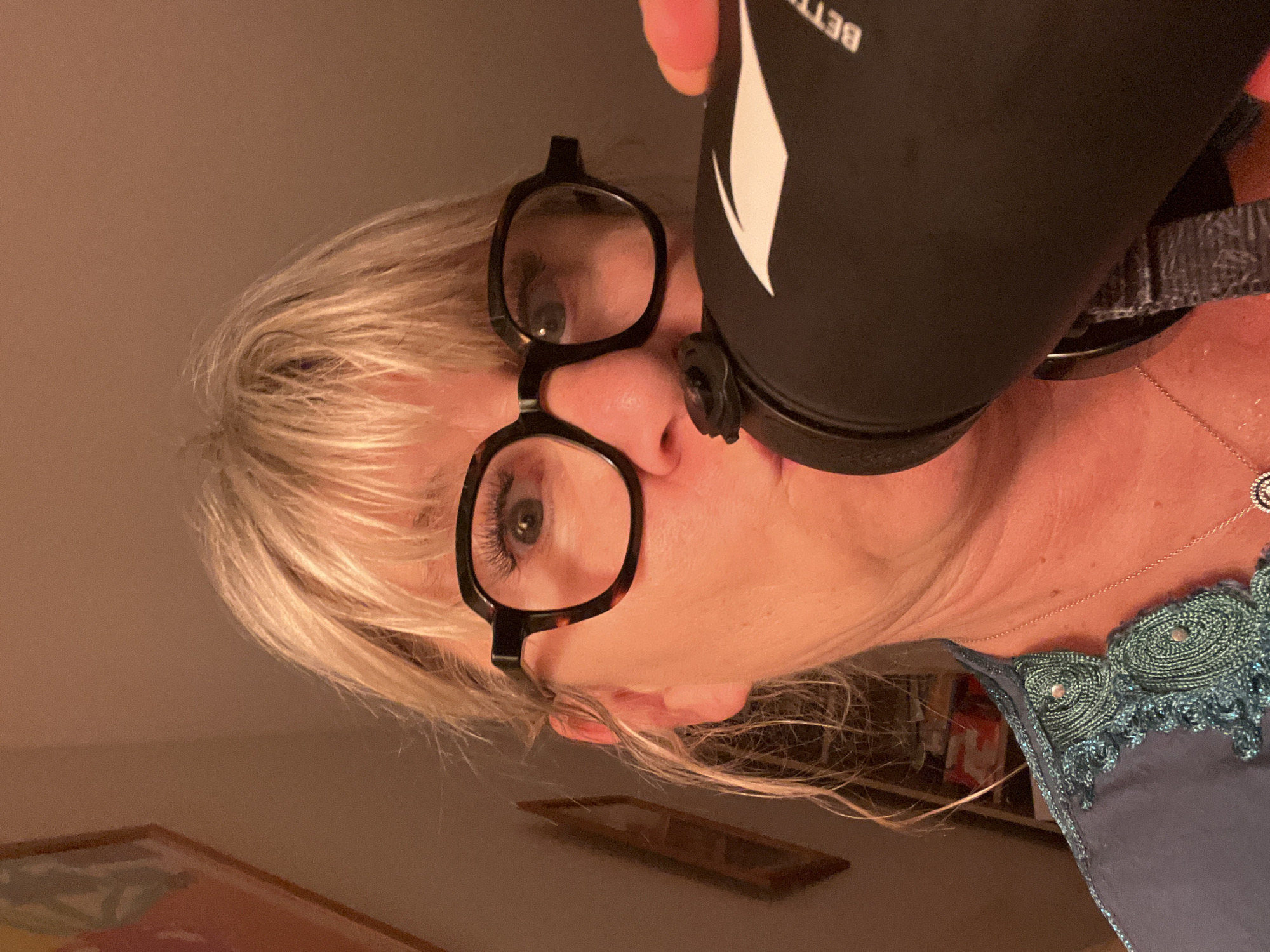 A fair-skinned woman with blonde hair is drinking from a black water bottle. She is wearing black-framed glasses. Her right hand is holding the bottle. She is wearing a dark blue shirt with teal crocheted detailing and a delicate silver necklace. The background is blurred but appears to be a room with neutral-toned walls and artwork hanging on them. The location, Chambœuf, France is not evident in the image.