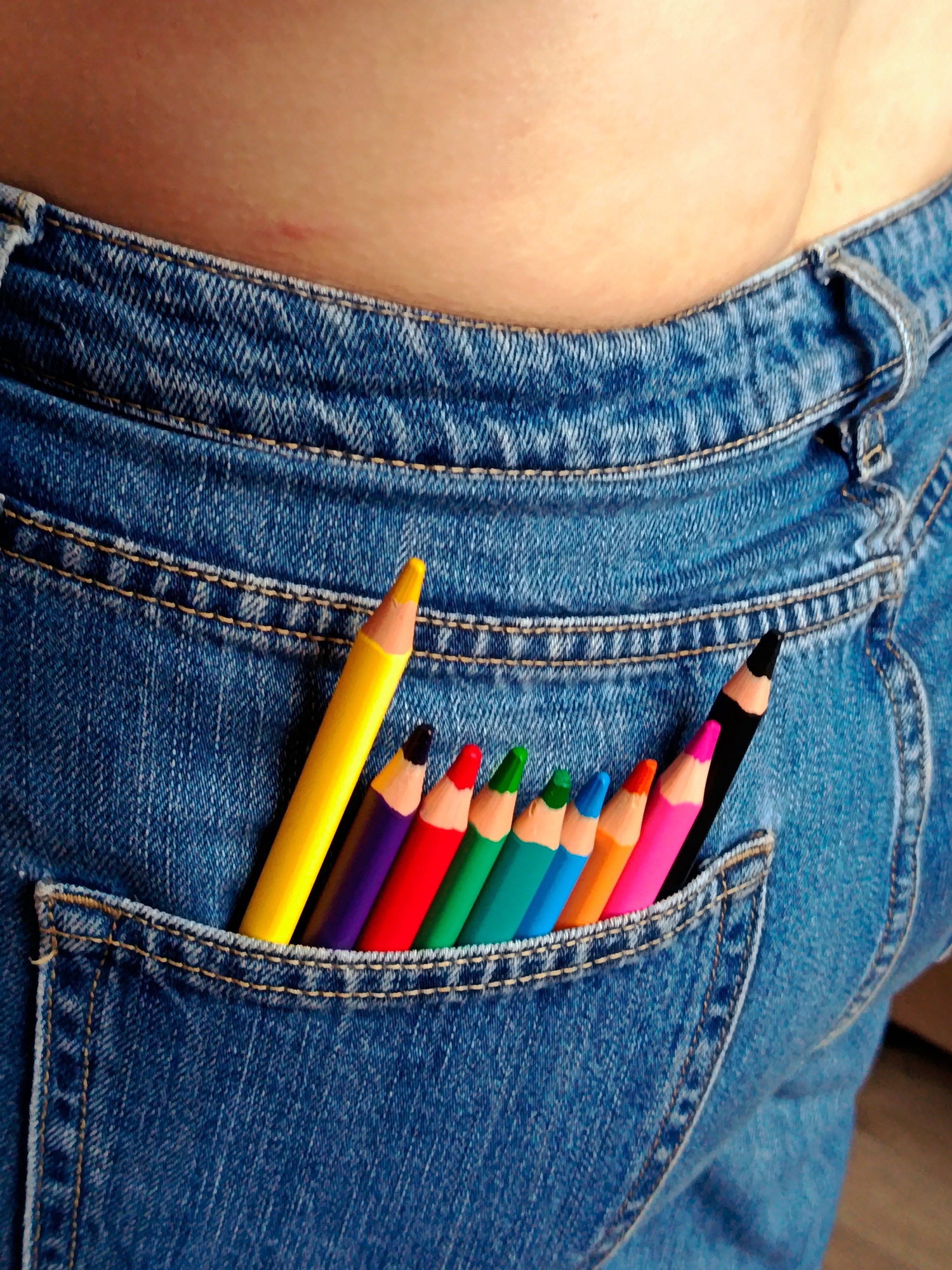 The image depicts a close-up shot of a person wearing blue jeans with a set of colored pencils sticking out of the back pocket. The pencils are arranged in a row, showcasing a variety of colors including yellow, purple, red, green, light blue, orange, pink, and black. A portion of the person's skin is visible above the waistband of the jeans. There is no indication of the location being in Фоки, Russia.