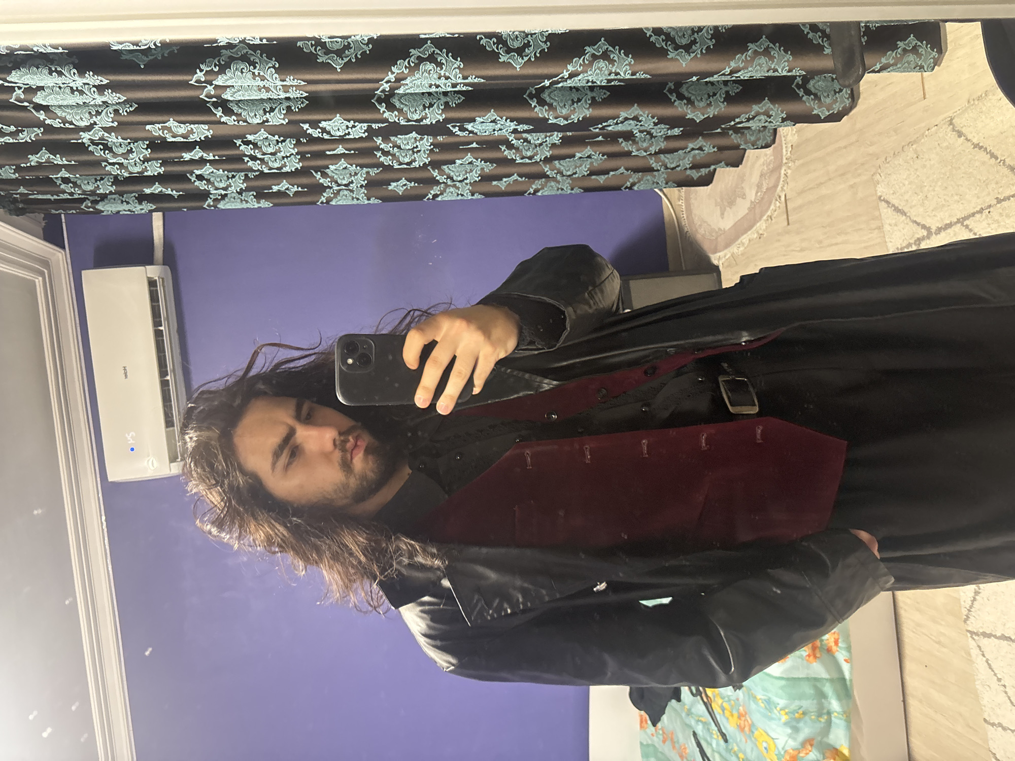 A young man with long, dark hair and a beard stands in an indoor setting, taking a mirror selfie. He is wearing a black leather trench coat over a black button-down shirt and a burgundy velvet vest. Black trousers and a black belt with a silver buckle are also visible. The background shows a purple wall with an air conditioning unit mounted near the ceiling. To the right, a patterned curtain hangs. To the left, a portion of a bed with a floral-patterned cover is visible, along with a rug on the floor.
