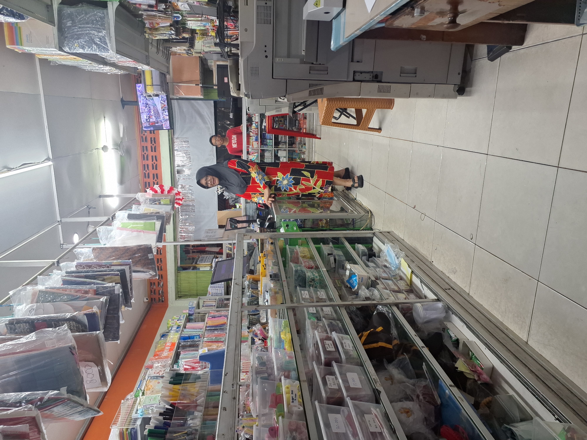 The image depicts the interior of a retail store in Balikpapan, Indonesia, likely a stationery or variety store. The store is densely packed with shelves filled with various products, including what appear to be stationery items, plastic containers, and other goods. A woman wearing a black hijab and a red and black patterned dress stands in the foreground, looking towards the left. Behind her, a young man wearing a red t-shirt with the word "NEVADA" printed on it is visible. In the background, a television screen displays an image, possibly showing store inventory or promotional content. Prominently featured in the right side of the image is a large office copier. The overall impression is of a well-stocked, functional retail environment.
