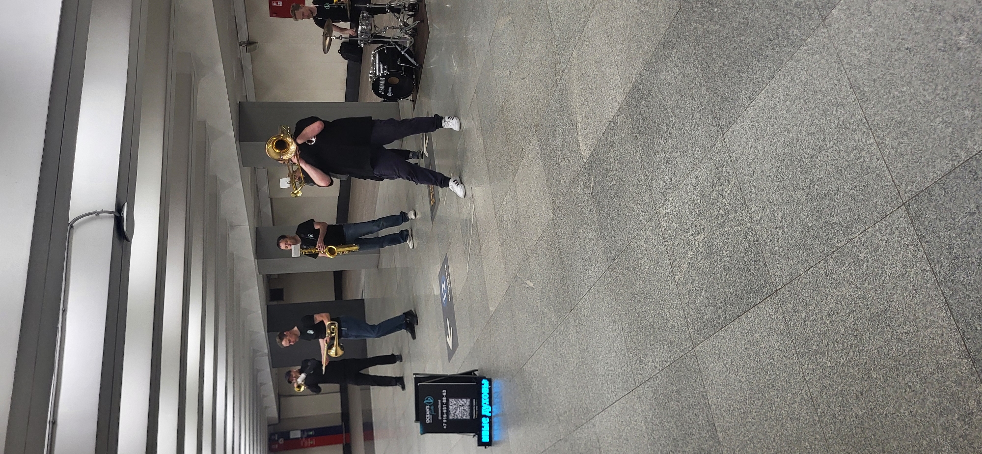 The image captures a group of four male musicians performing or practicing in a spacious indoor setting, likely an underground passage or subway station. The floor is covered with light gray, speckled granite tiles arranged in a diamond pattern. The walls and ceiling feature a modern design with alternating vertical panels of light and dark gray, illuminated by recessed artificial lighting.
In the foreground, a man wearing a black t-shirt, dark trousers, and white athletic shoes plays a trombone. Behind him, another man in a dark t-shirt and jeans plays a saxophone. Further back, a third man, also in a dark t-shirt and jeans, plays a trumpet. In the background, a fourth musician is seated behind a black drum set, which has "SONOR" visible on the bass drum and "YAMAHA" partially visible on other components. All musicians are dressed in dark, casual attire.
On the right side of the image, a black digital display board with blue glowing text is visible. It shows "OCEANS", a phone number "+7 916-651-88-93", a QR code, and the Cyrillic text "ДЖАЗ В ДУХОВЫЕ" (Jazz in Brass/Wind instruments). A blue directional arrow sticker is also visible on the tiled floor near the sign. The scene depicts a musical performance or rehearsal in a public, brightly lit space.