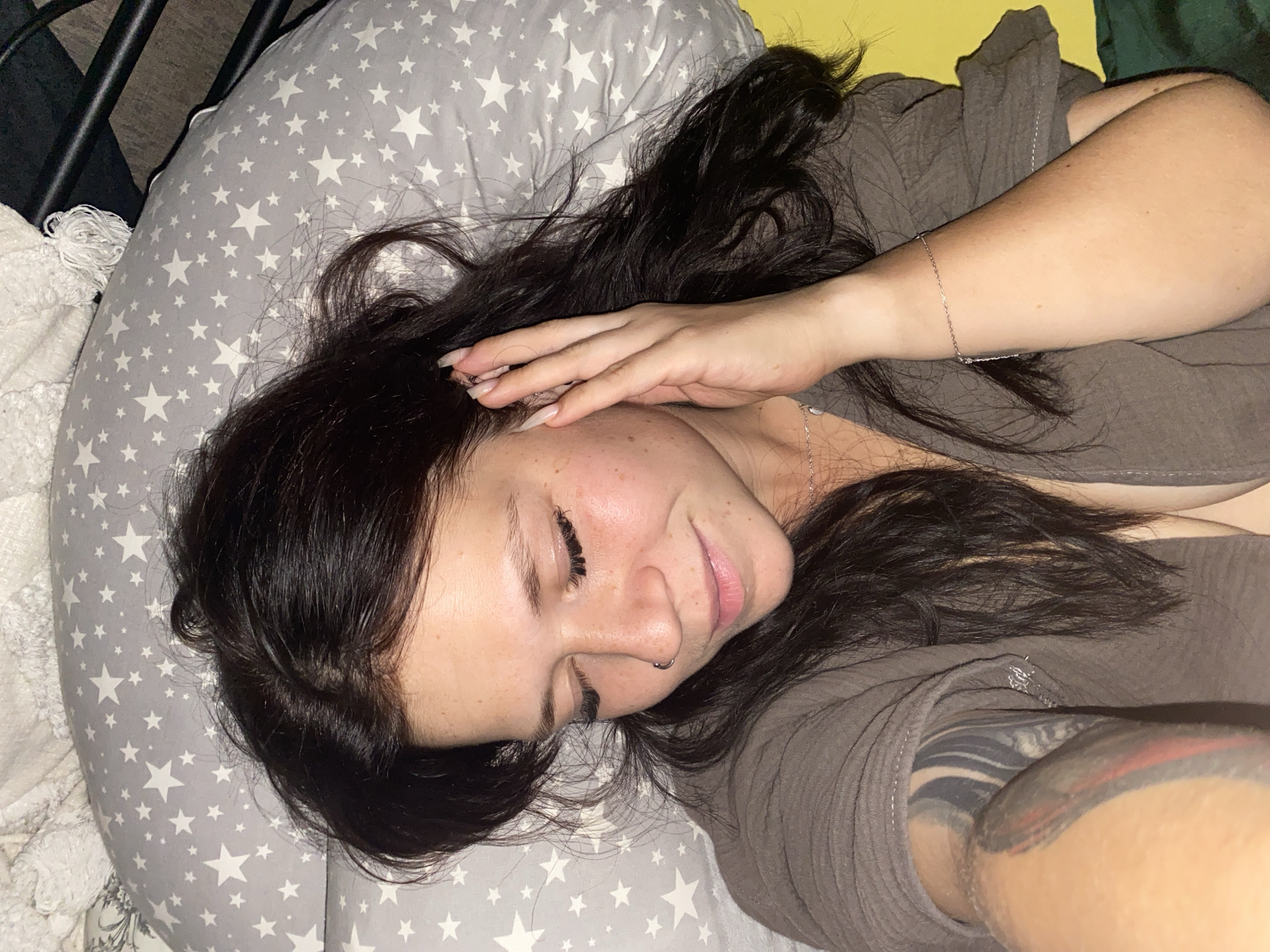 A woman with dark hair and freckles lies on her side, resting her head on a grey pillow patterned with white stars. Her eyes are closed, and her hand is raised to her ear. She is wearing a brown shirt and a necklace, and has a nose ring and a tattoo on her arm. The background is out of focus, with hints of yellow and green. The scene appears to be in a bedroom.