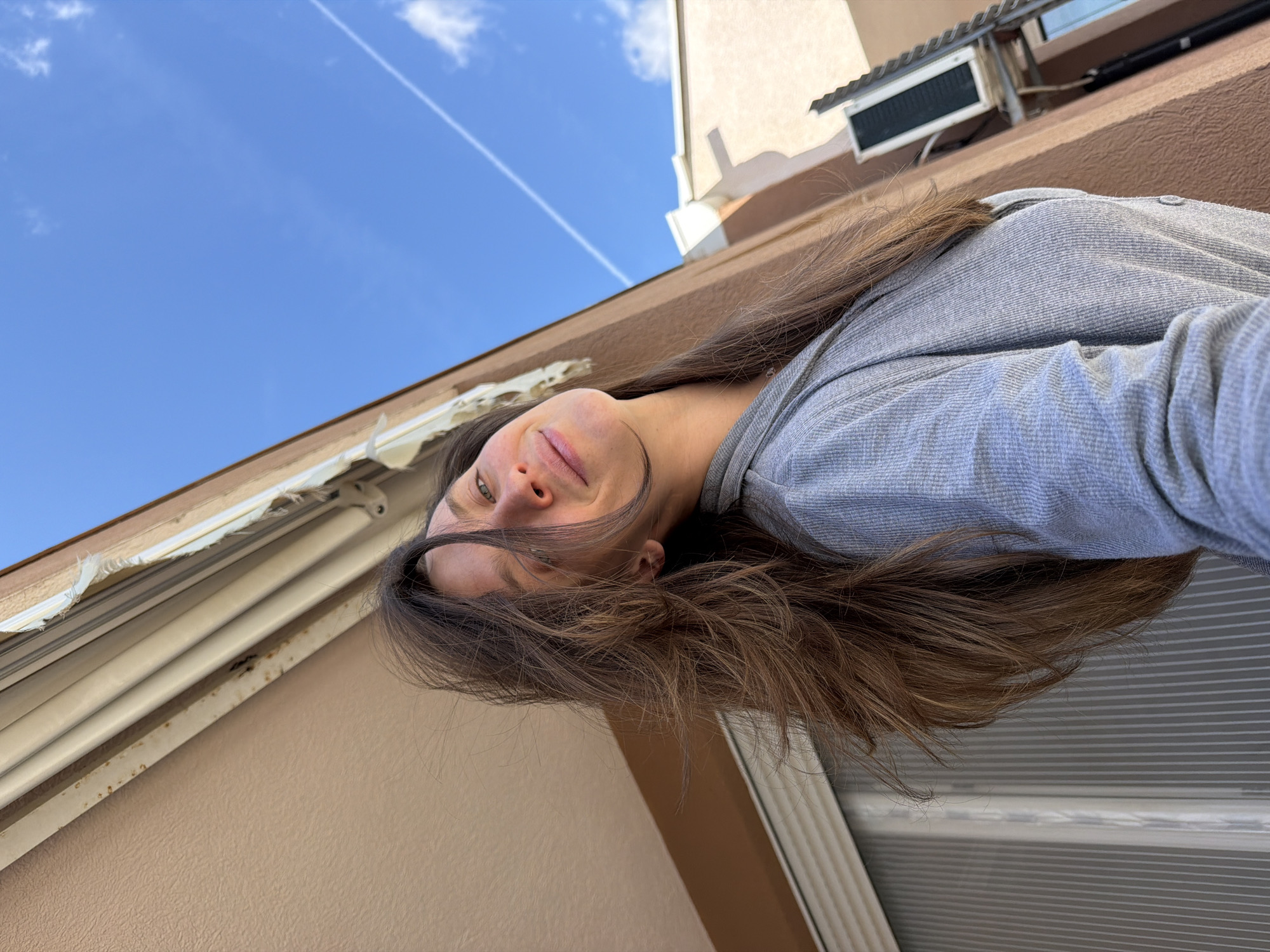 A woman with long brown hair and green eyes is looking at the camera. She is wearing a gray long-sleeved shirt. The background shows a building, blue sky, and a contrail. It is impossible to identify the location as Budva, Montenegro, with the provided information.