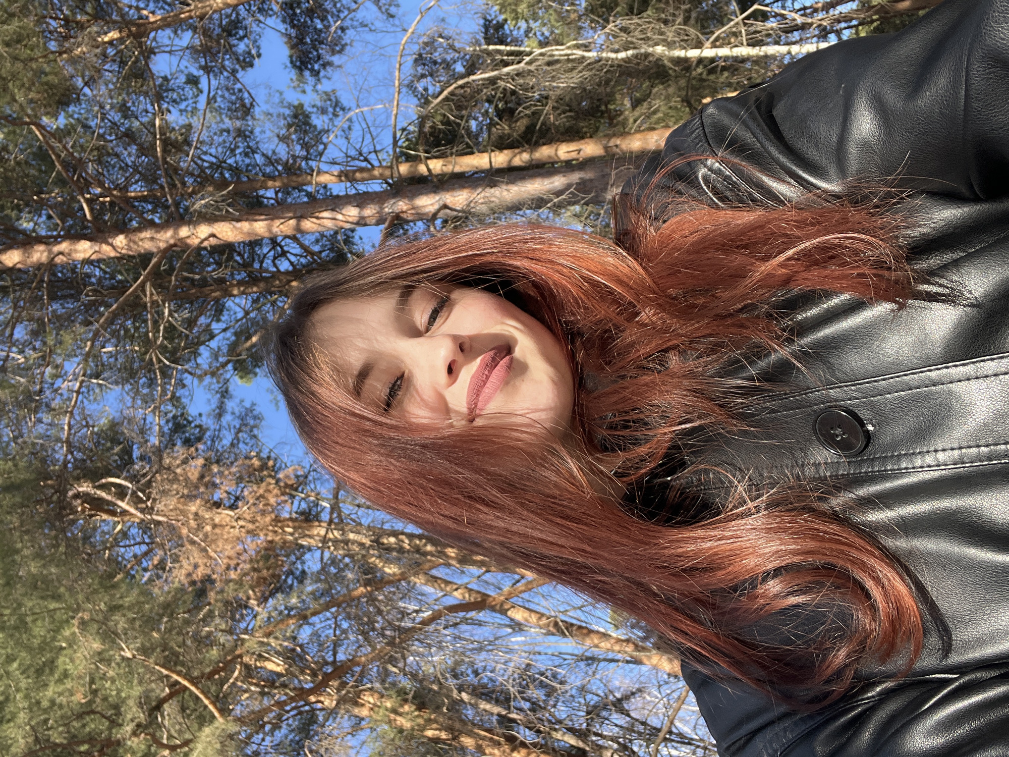 A young female, with long, reddish-brown hair and wearing a black faux leather jacket, is depicted in a close-up, selfie-style photograph, smiling faintly. Her face is illuminated by direct sunlight, creating subtle shadows. She is positioned outdoors under a clear blue sky. The background consists of numerous tall trees, including pine trees with green needles and deciduous trees with bare branches, suggesting a wooded area. This scene is located in a forested or park area within the city of Chaikovsky, Russia.