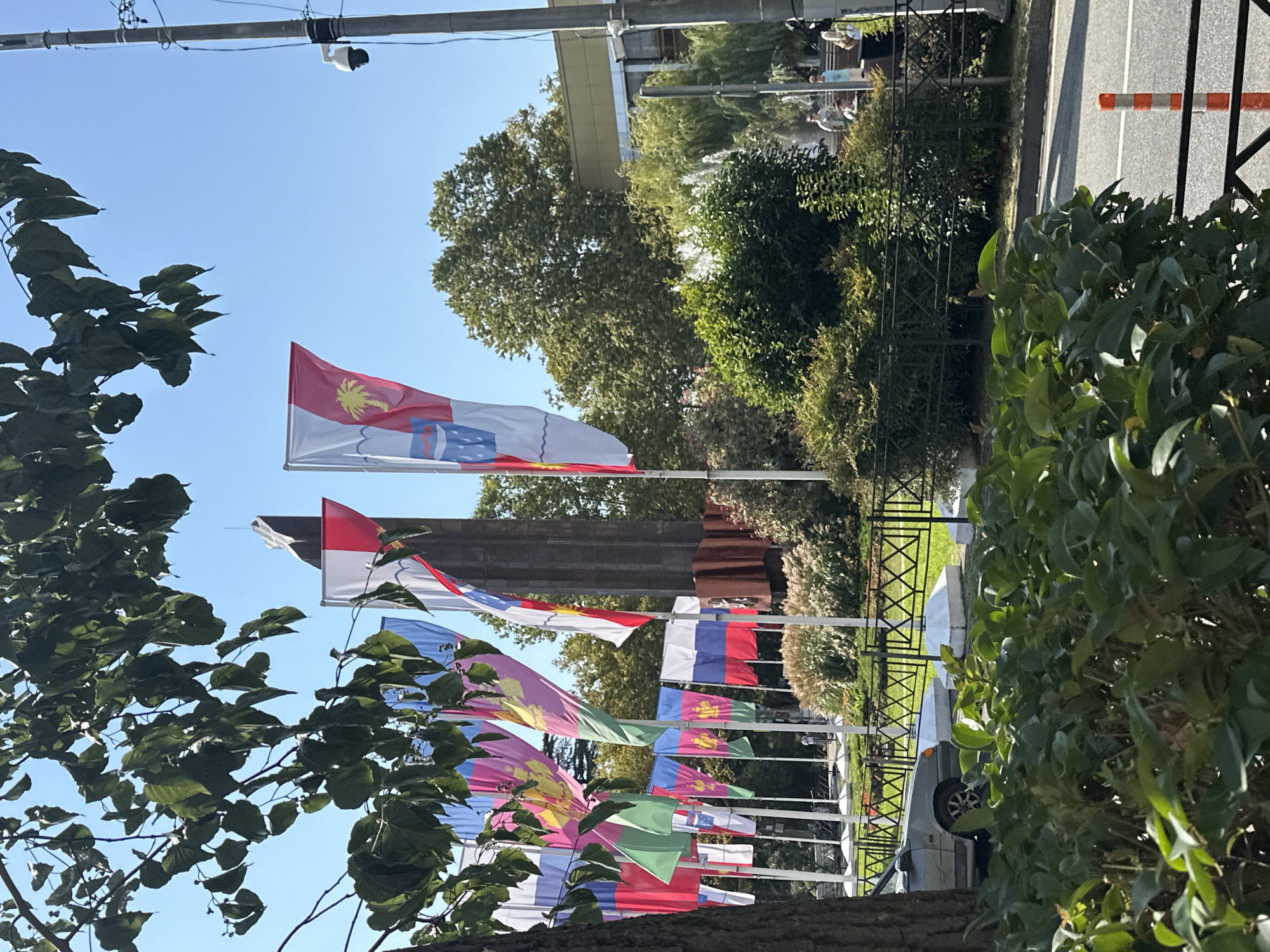 This image depicts a sunny day in Sochi, Russia, likely in a public area such as a park or promenade. In the foreground, lush green bushes partially obscure a black metal fence. Beyond the fence, a grey sedan is parked on a paved surface. A line of flags, predominantly red, white, and blue, are displayed on poles, some featuring intricate designs and symbols. Behind the flags, a tall, dark monument stands prominently against a clear blue sky. Trees with dense green foliage are visible in the mid-ground, and a water fountain with a white spray is noticeable amongst the greenery. A few individuals are present in the background, appearing to be walking or standing around. A security camera is mounted on a pole on the right side of the frame, indicating a public or monitored space. The overall scene suggests a civic or memorial space within the city of Sochi.