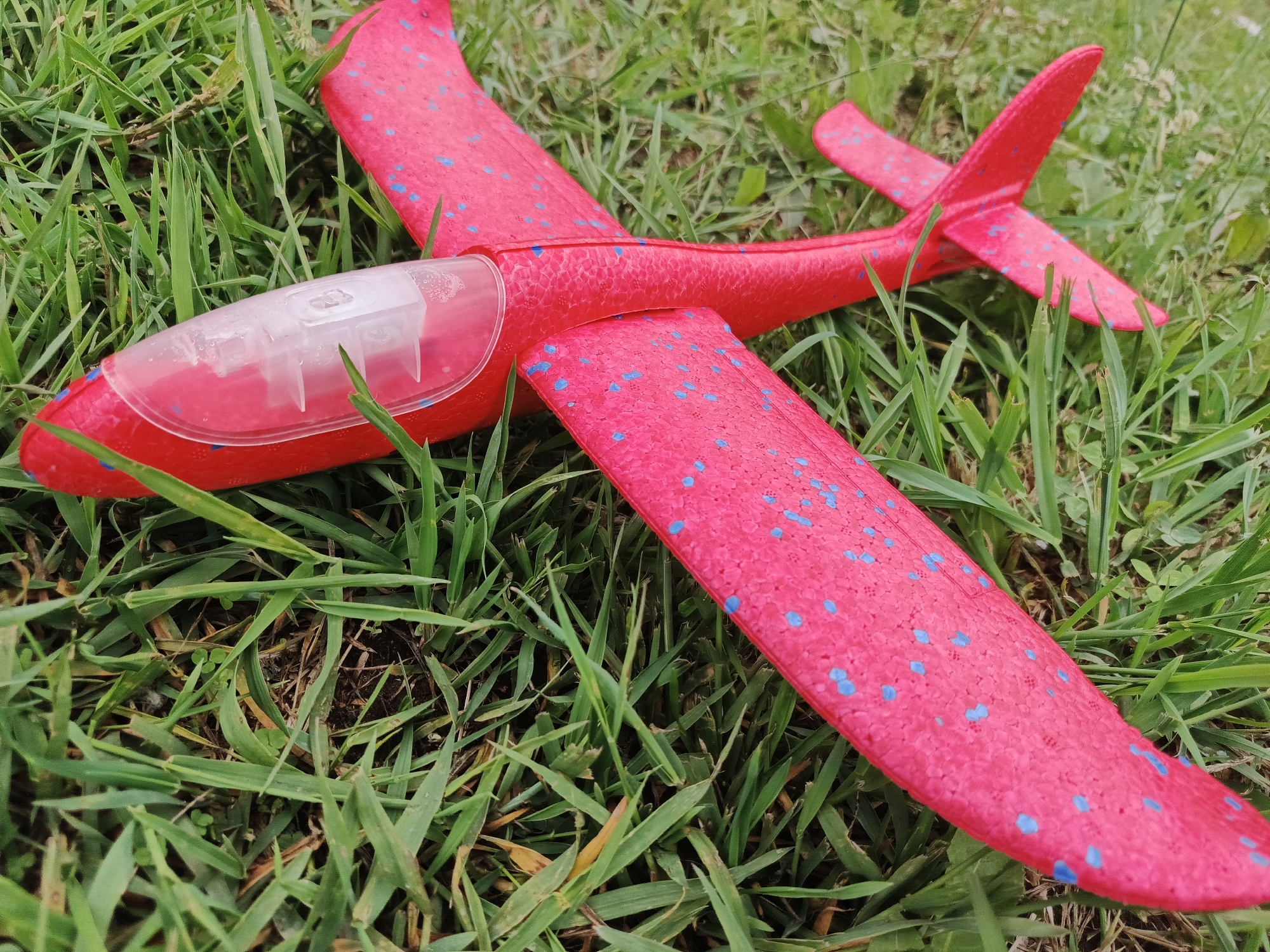 A bright pink foam toy airplane with scattered blue speckles is positioned on a dense patch of green grass. The airplane features a main wing, a slender fuselage, and a T-tail configuration, where the horizontal stabilizer is mounted atop the vertical stabilizer. A translucent, clear plastic canopy-like component is affixed to the nose of the fuselage, revealing white internal structural elements. The foam material of the airplane exhibits a textured, porous surface. The grass in which the airplane rests is predominantly green, with blades varying in length and density, interspersed with some dried brown blades and small, broad-leafed ground cover plants. Dark brown soil is visible in sparse patches beneath the grass. The scene is illuminated by natural, diffuse daylight. No individuals or animals are present. The airplane is stationary on the ground. The location is in the city of Unknown, Unknown.