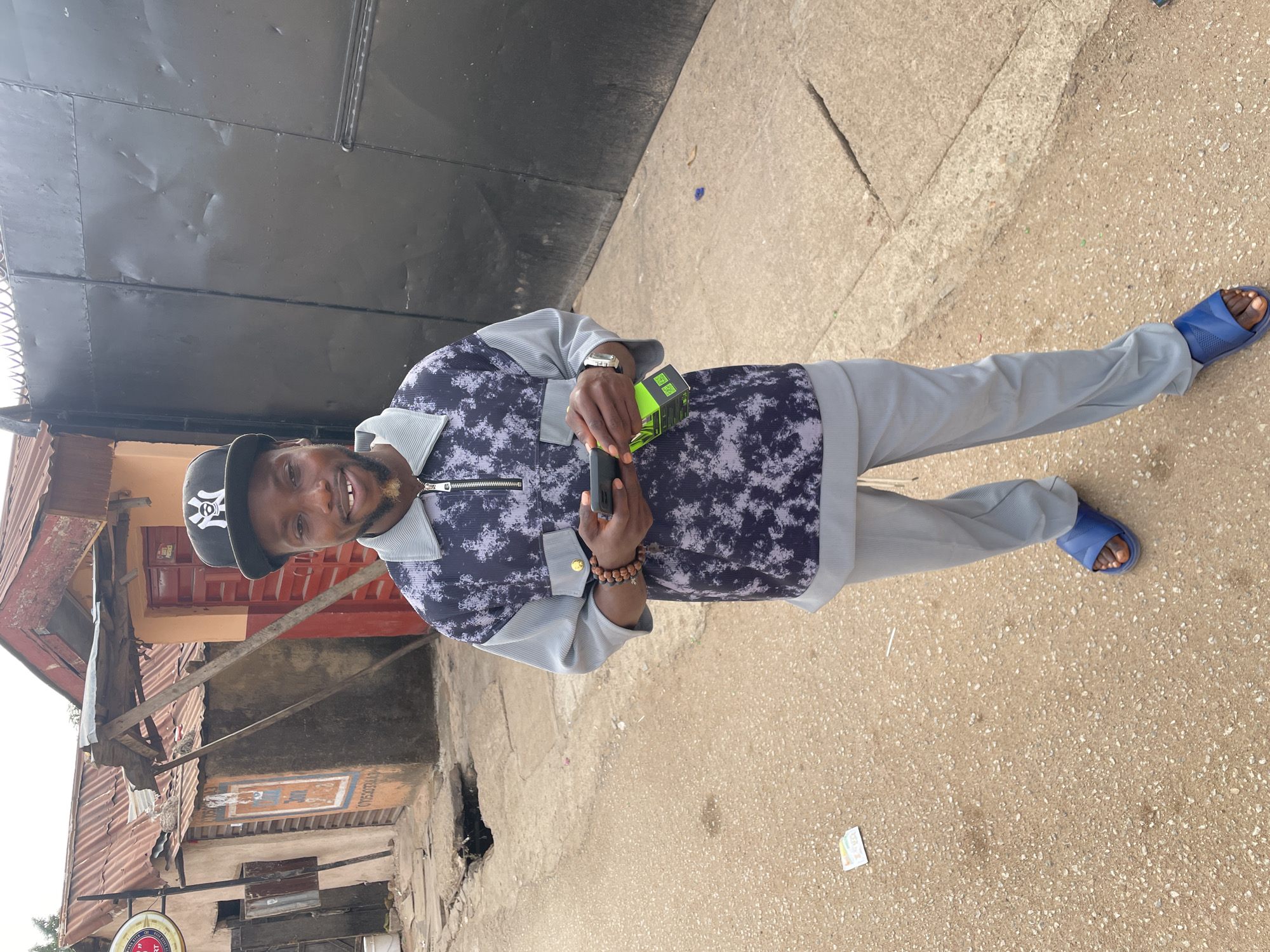 A man stands on a sidewalk near a building, holding a small box and a black object in his hands. The man wears a patterned top with light gray sleeves and pants, a baseball cap, blue sandals, and a wristwatch. He is smiling. The sidewalk is light brown and textured. Behind him are buildings with aged facades, suggesting an older part of the city. A large metal door or gate is also visible in the background. The man appears to be posing for the photo. This location is in Jos, Nigeria.