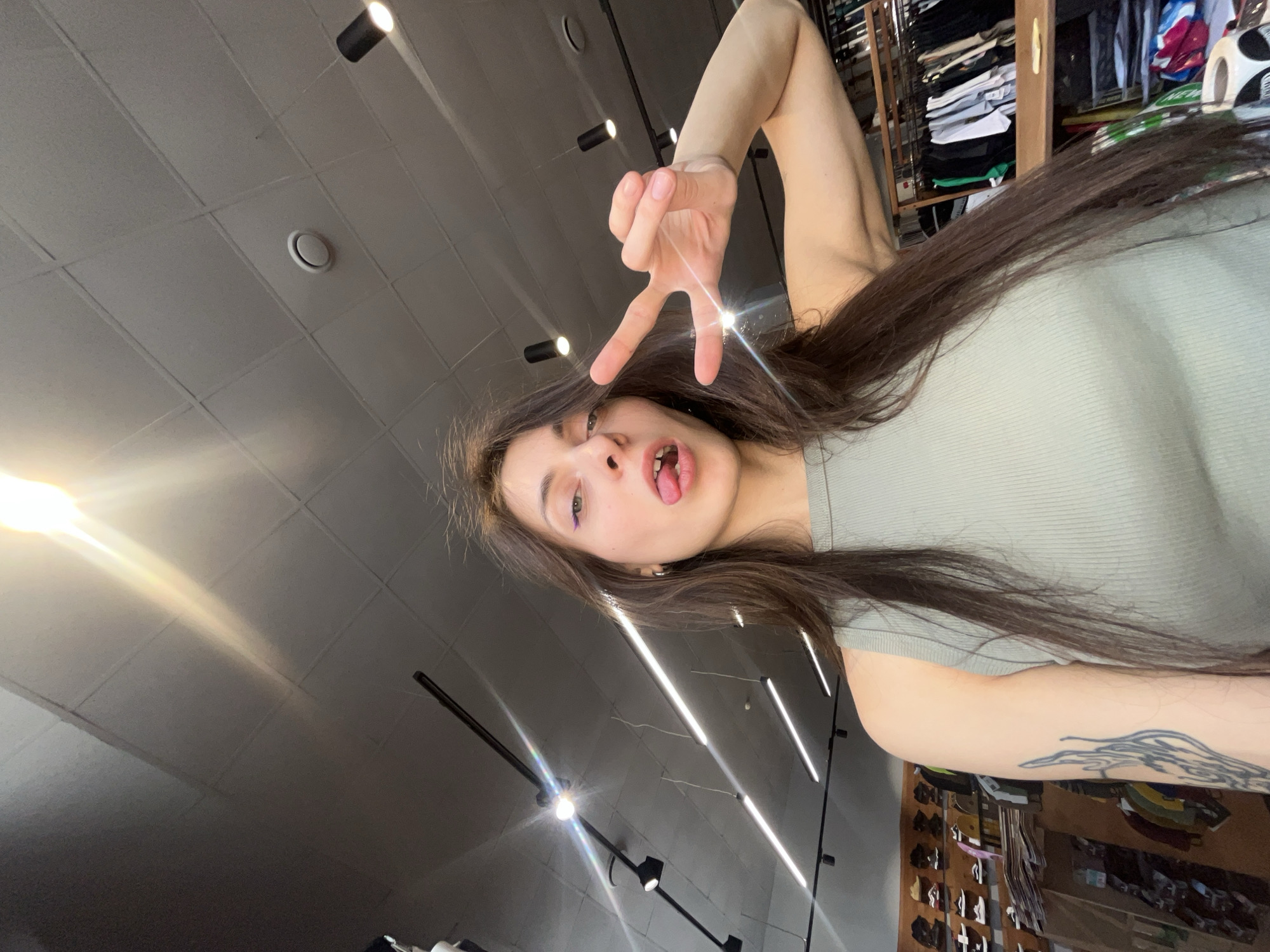 A young woman is taking a selfie, holding up a peace sign with her right hand and sticking out her tongue. She is wearing a light green top and has long, dark brown hair. The selfie is taken from a low angle, looking up towards a gray, tiled ceiling with track lighting. In the background, there are racks of clothing and shoes, suggesting she is in a retail store. Based on available information, the location of this scene is unknown in Krasnoyarsk, Russia.