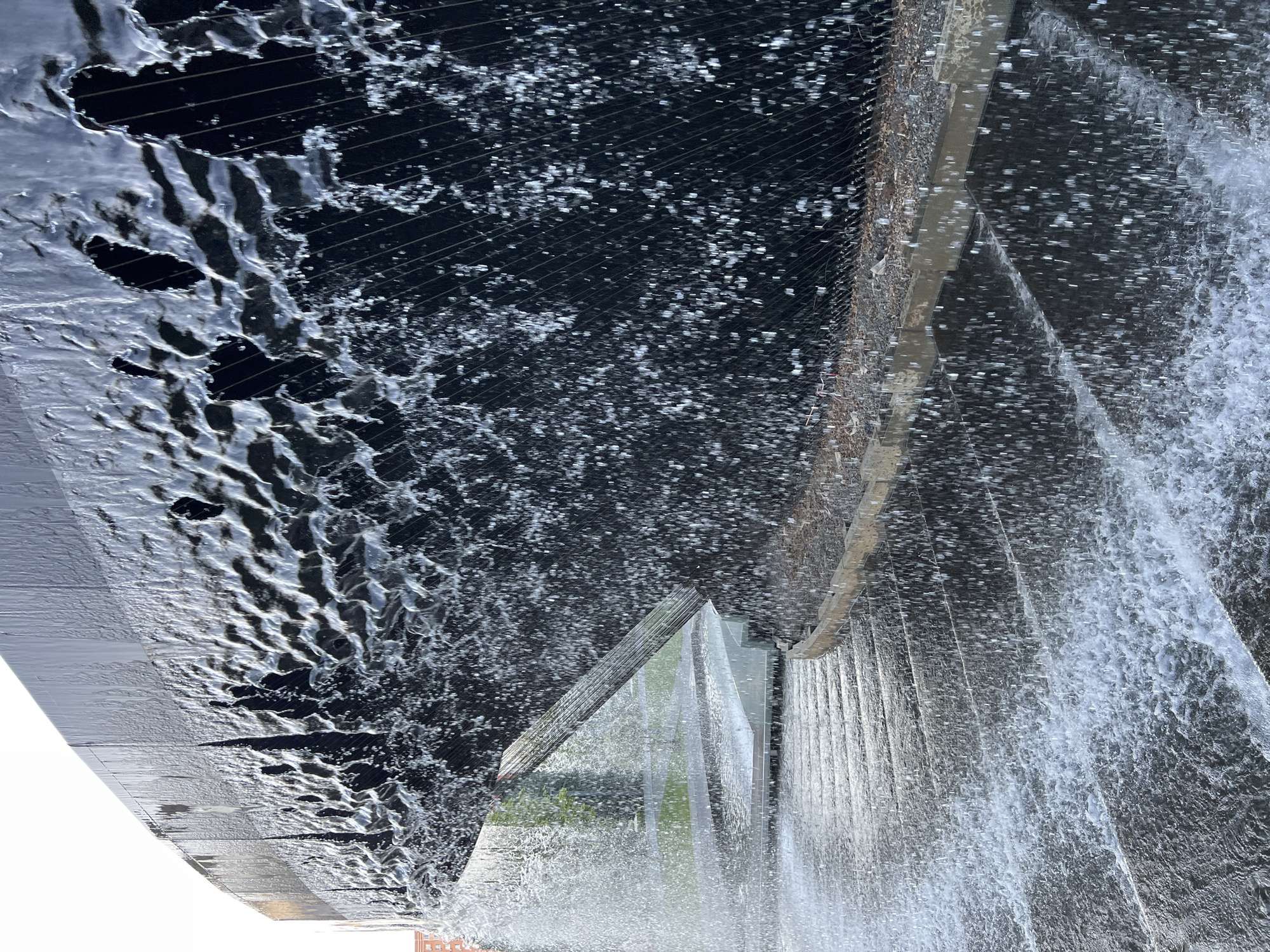 The image shows a close-up of a dark, curved surface with water flowing over it and cascading down a tiered, textured surface. The dark surface, likely part of a building or monument, has irregular openings and appears to be made of metal. Thin, vertical lines are visible on its surface, possibly a mesh or grating. Water streams from these openings, creating a dynamic waterfall effect. Below, the tiered surface is wet and reflects the water. A light-colored edge, possibly made of wood or stone, runs along the top of the tiered structure. In the background, a hint of green foliage and a building facade are visible. The scene suggests an outdoor fountain or architectural water feature. This location is in Krasnodar, Russia.