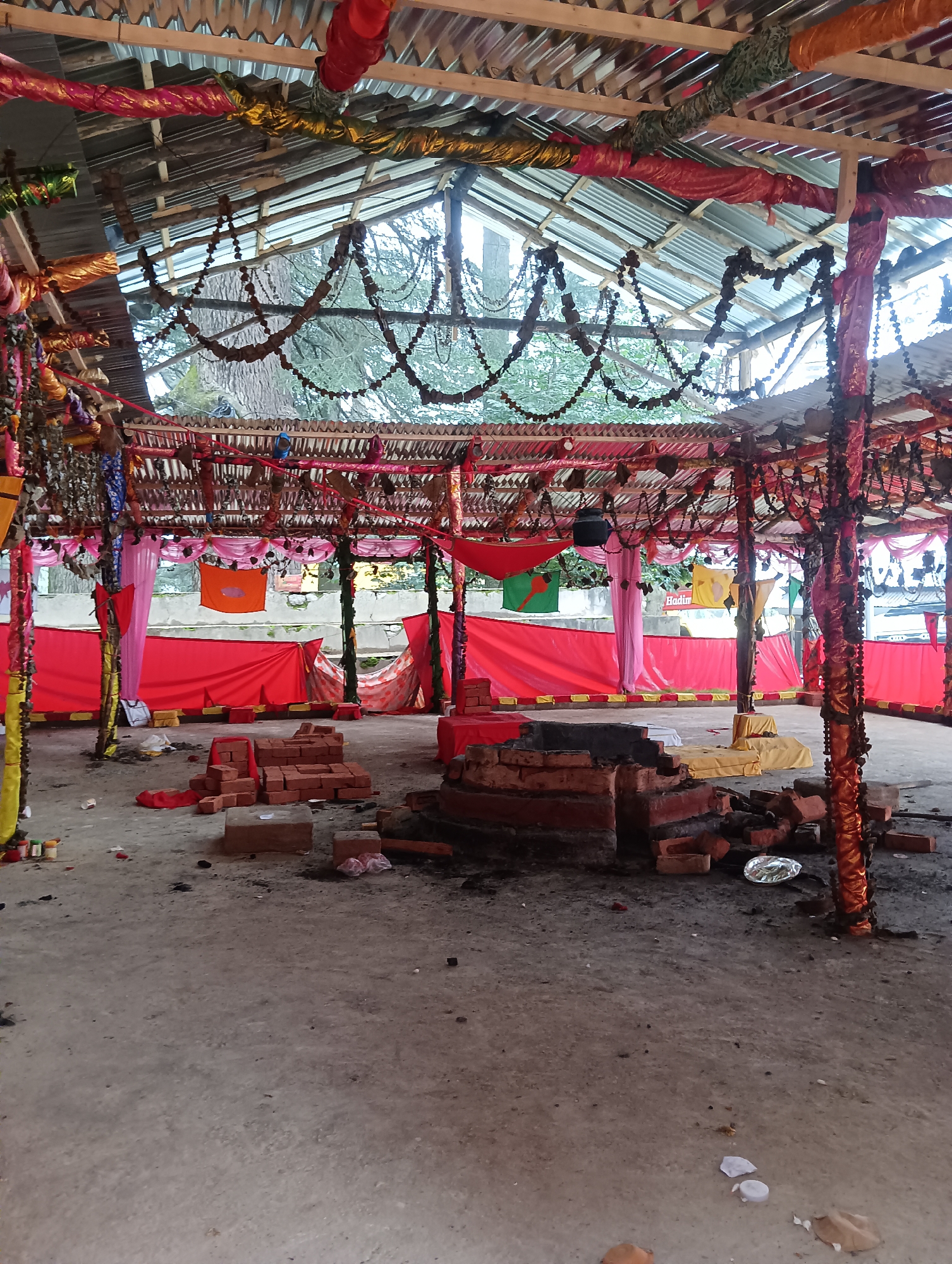 The image depicts an outdoor, open-air structure with a corrugated metal roof supported by wooden beams and columns. The structure is adorned with festive decorations, including colorful draped fabrics in red, pink, yellow, and orange, as well as beaded garlands hanging from the ceiling. Several flags and banners are also visible, some with what appear to be Bengali text. The ground is made of concrete and shows signs of wear and debris, including scattered bricks, paper wrappers, and plastic items. In the foreground, there are piles of bricks, suggesting construction or renovation work, and a brick structure that resembles a makeshift altar or fire pit. The overall atmosphere is one of a temporary ceremonial or event space, possibly in a rural or semi-urban setting. The mention of Manali, India, suggests the possibility that this is a Hindu temple or a place of religious gathering, although no specific religious iconography is clearly identifiable. The scene appears to be in a state of disarray, possibly after an event or during preparation.