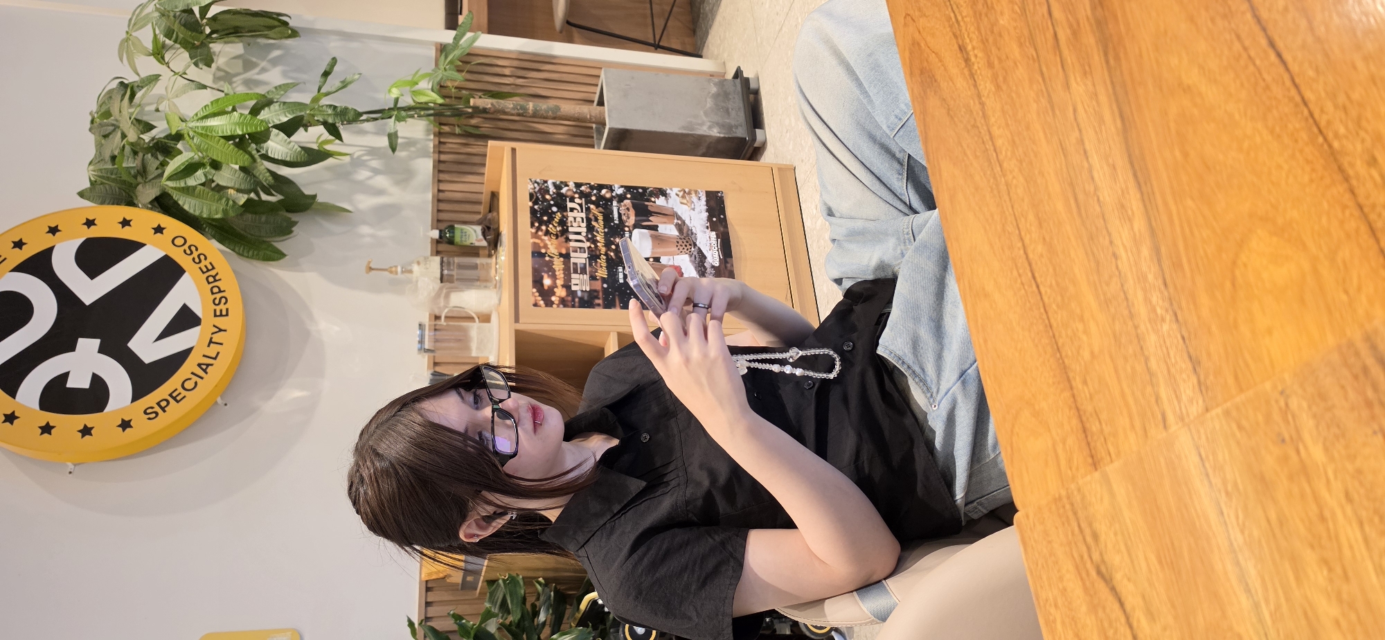 In a brightly lit interior, a young woman with brown hair pulled back in a ponytail and wearing black-rimmed glasses is seated in a chair. She is looking down at her mobile phone, which she holds in her hands. She is dressed in a black collared shirt and light blue denim pants. A string of white beads hangs from her hand. In the background, a wooden cabinet displays various items including a poster advertising drinks. A tall potted plant is situated to the right of the cabinet. Above the cabinet, a circular yellow sign with black lettering and stars is visible. The scene appears to be a cafe or a similar establishment. This location is in Cheongju-si, South Korea.