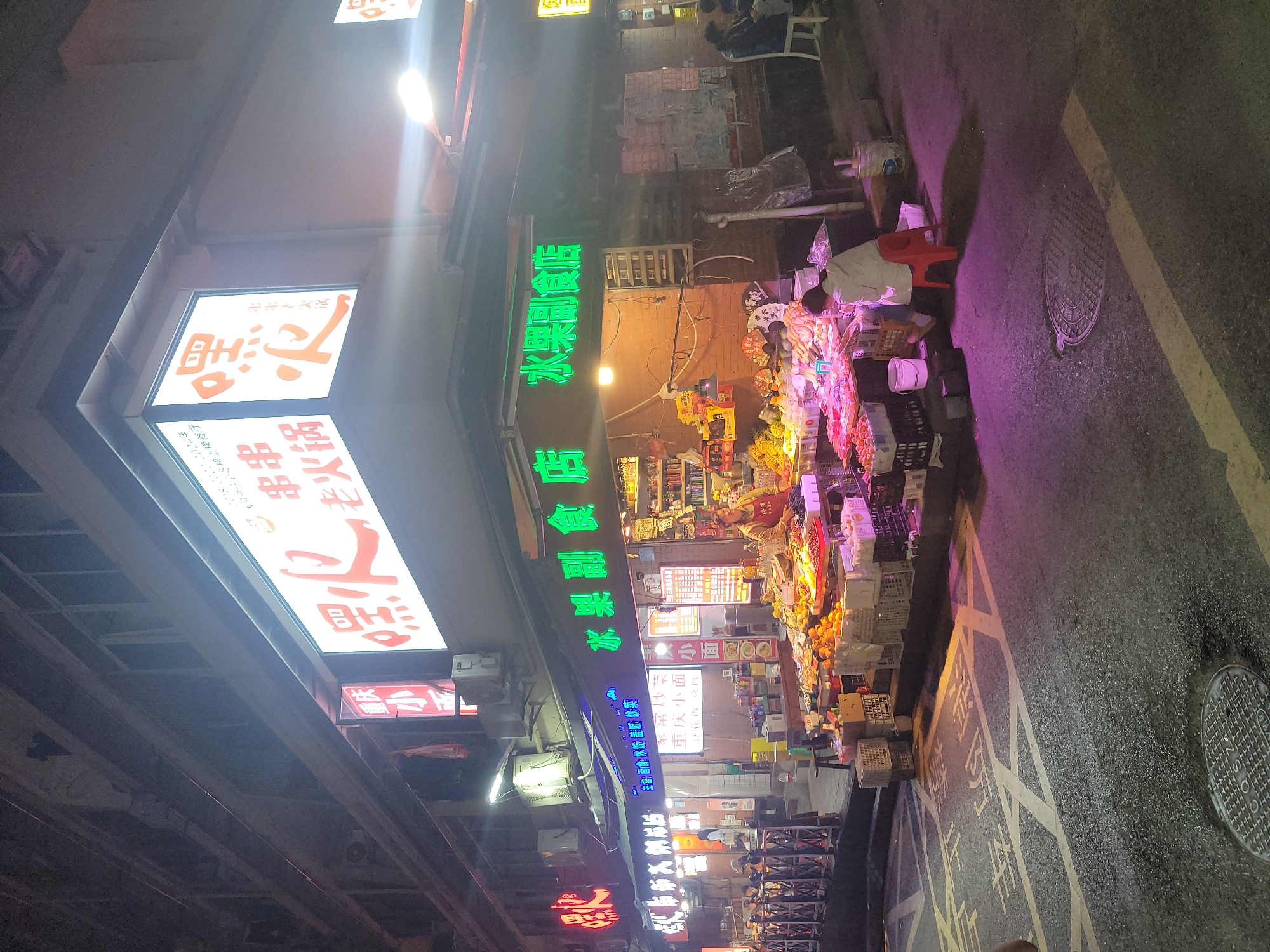 This media file captures a vibrant night scene on a bustling street in Yuzhong District, China. The setting is an urban night market or food street, brightly illuminated by numerous neon signs and glowing lightboxes.
Several people are visible, engaged in various activities. A vendor is seen tending to a street stall laden with produce, while another person sits on a red plastic stool. Other individuals are present in the background, likely shoppers or passersby.
The foreground is dominated by various market stalls displaying an abundance of fresh fruits and vegetables in crates and baskets. The street itself is asphalt with white painted markings and a visible manhole cover. Above the street, large building structures or an elevated roadway are present.
The scene is characterized by a multitude of illuminated Chinese characters. Prominent green neon signs advertise "永常炒" (Yongchang Stir-fry) and list various dishes such as "主食面食炒饭盖饭炒菜" (Staple food, noodles, fried rice, covered rice, stir-fry). Other bright signs display "老火锅" (Old Hotpot), "串串" (Skewers), and "陽上場餐厅" (Yangshangchang Restaurant). Large white signs repeatedly feature "水果副食店" (Fruit and grocery store), suggesting a prominent produce vendor. Smaller signs mention "重庆小面" (Chongqing small noodles), "苏小王" (Su Xiaowang), and "以花改日," contributing to the lively commercial ambiance of the night market.