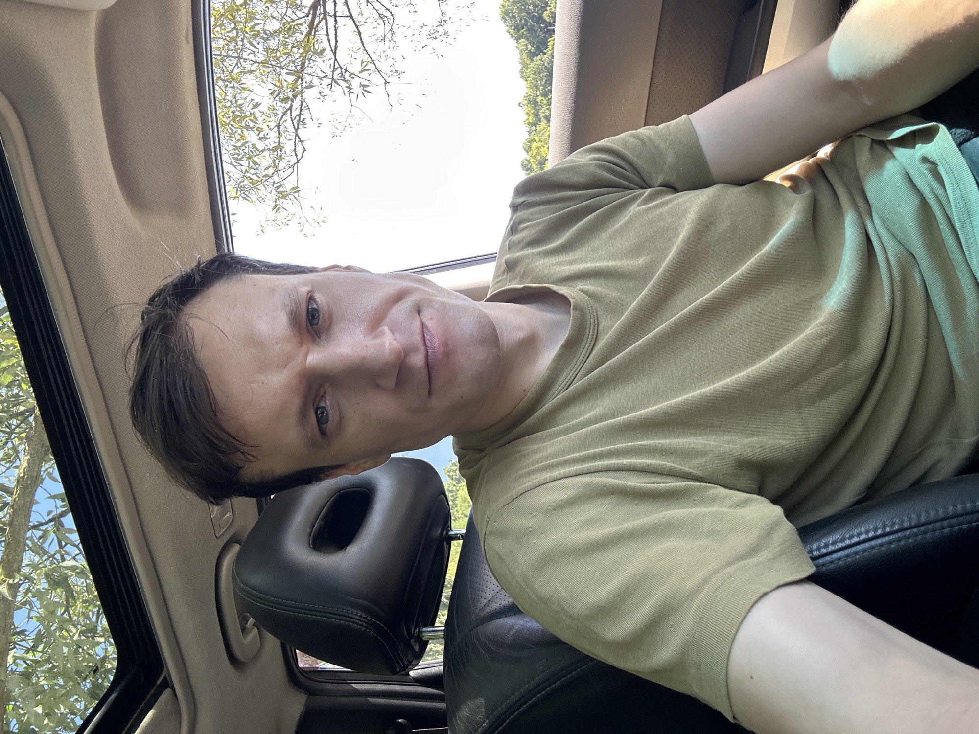 A man with short, dark brown hair is sitting in the passenger seat of a car, looking directly at the camera. He is wearing a short-sleeved, olive green t-shirt. The car has a sunroof, which is open, revealing a tree and blue sky above. The interior of the car is visible, including a black leather headrest and seat. The background shows a landscape with trees and foliage, suggesting an outdoor setting, possibly in a rural area or on the outskirts of a city like Makiv, Ukraine, given the natural light and vegetation. The man's expression is neutral, with a slight smile.