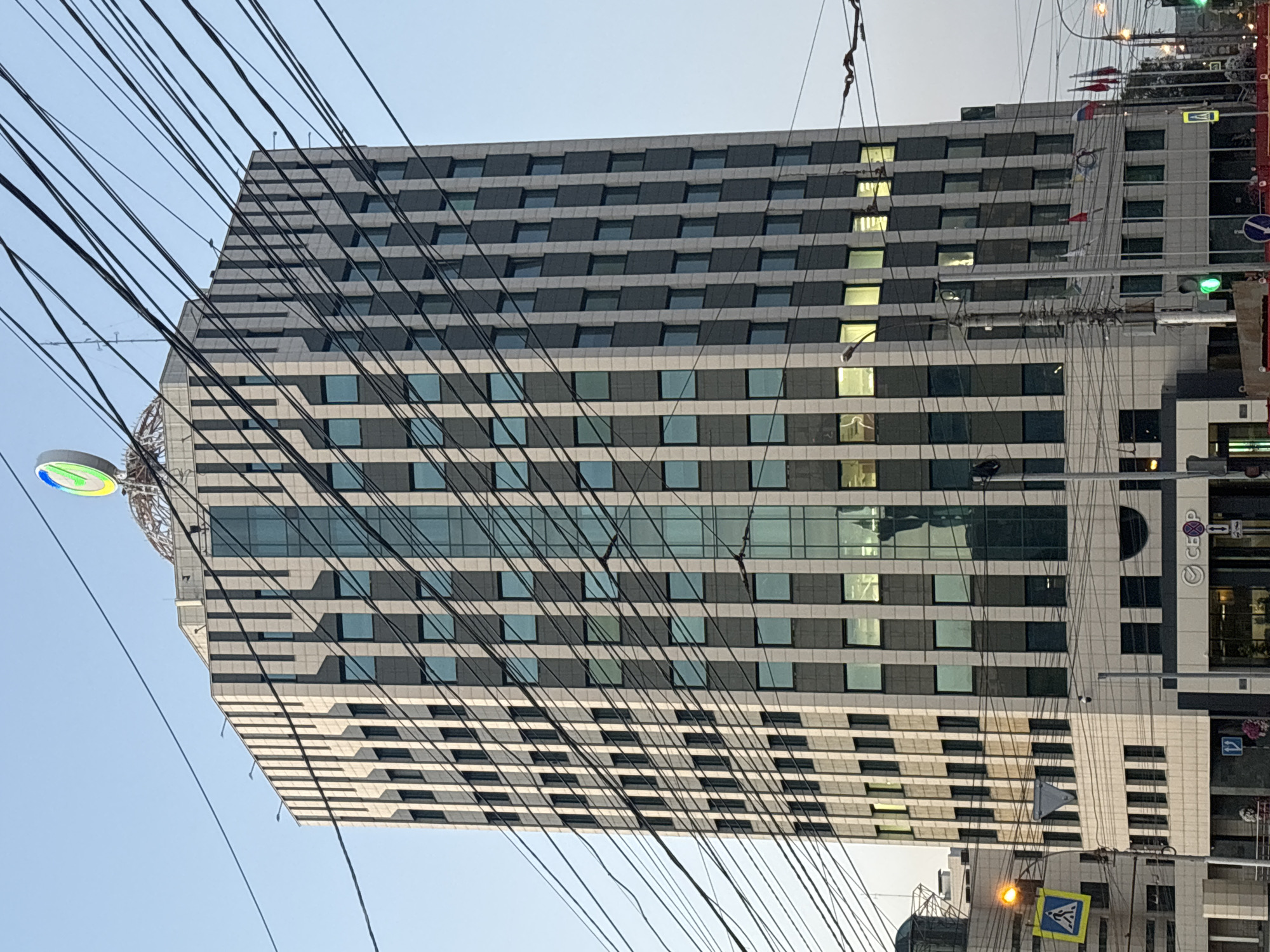A multi-story office building with a light-colored facade and dark-paneled windows stands against a clear blue sky. A circular, green and yellow logo adorns the top of the building. Numerous black electrical wires crisscross the foreground, obscuring parts of the building. The base of the building features a sign with Russian text and a circular logo. Various flags are visible near the lower levels of the building. Street lights and traffic signals are also present, along with pedestrian crossing signs.