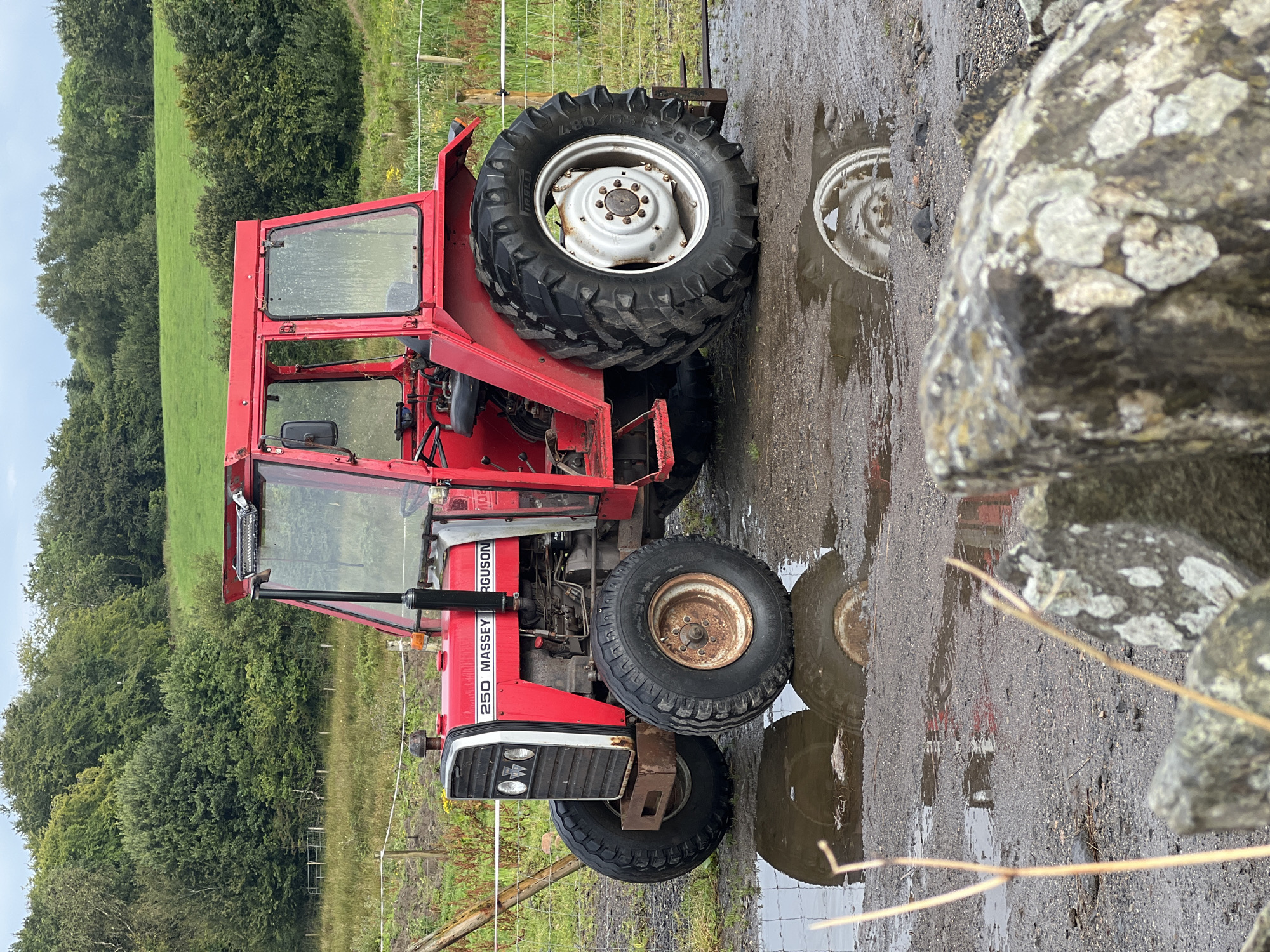 A red Massey Ferguson 250 tractor is parked on a muddy surface, with puddles reflecting the vehicle. The tractor is in a rural setting, with trees and a green field in the background. A wire fence runs horizontally across the image. In the foreground, there are large rocks and dry vegetation. The sky is overcast. The scene appears to be in or near Mountcharles, Ireland. There are no people visible in the image.