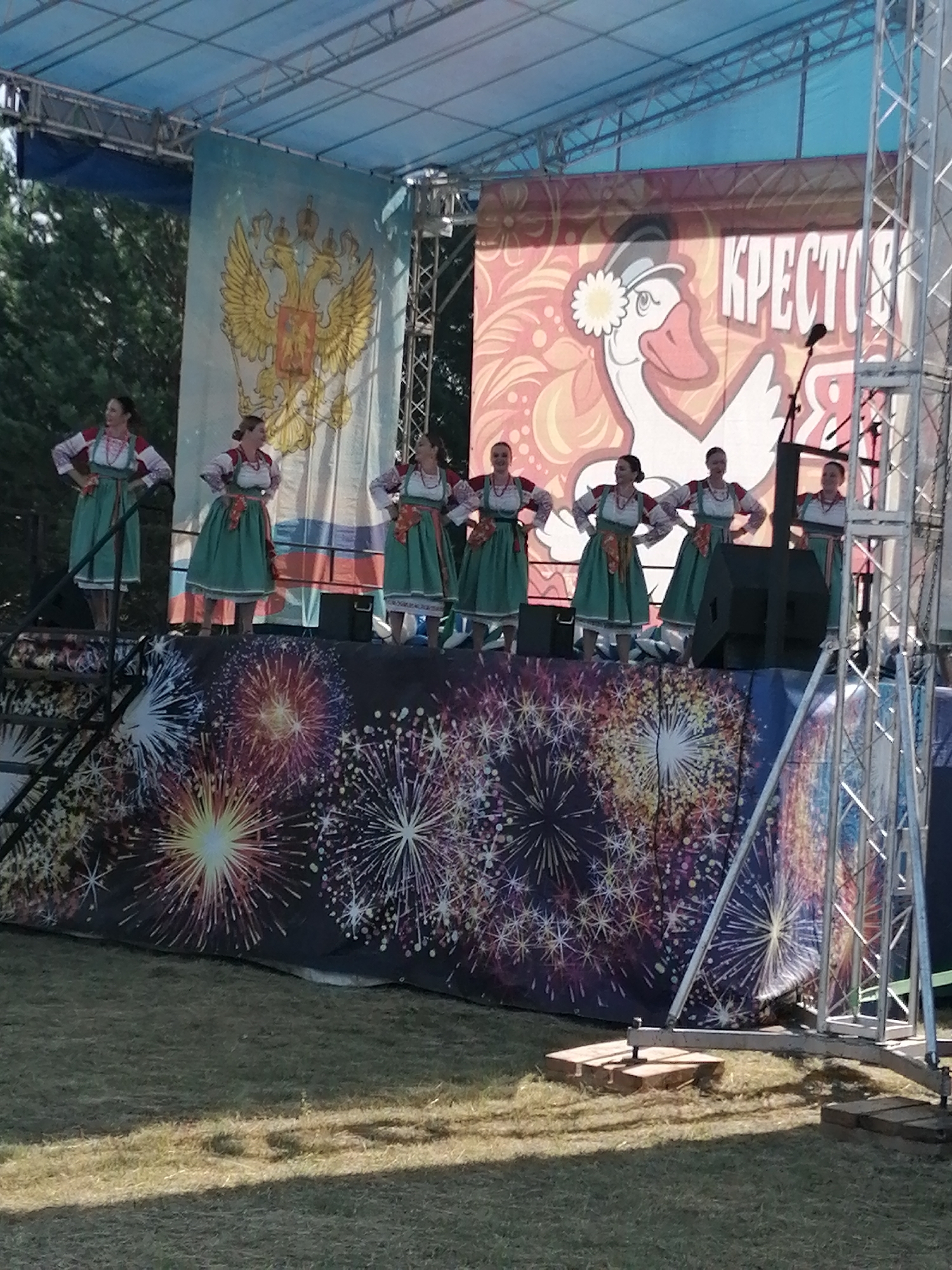 The image shows a performance stage set up outdoors, likely for a public event. On the stage, six women are dressed in traditional Russian folk costumes, which consist of embroidered blouses, sarafans (dresses), and aprons. They appear to be posing or performing a dance.
Behind the performers, there are several large banners. The one on the left features a prominent golden double-headed eagle, a symbol of Russia, against a light blue background. The banner in the center depicts a white goose wearing a cap adorned with a flower, set against a vibrant, patterned backdrop with the Russian word "КРЕСТЬЯНКА" (Krestyanka, meaning peasant woman) written in large white letters.
The stage itself is decorated with a backdrop featuring a colorful fireworks display. In front of the stage, the ground is covered with grass, and there are some structural elements of the stage setup visible, including metal scaffolding and wooden supports. The overall scene suggests a festive occasion, possibly a cultural festival or celebration in the city of Shadrinsk, Russia.
