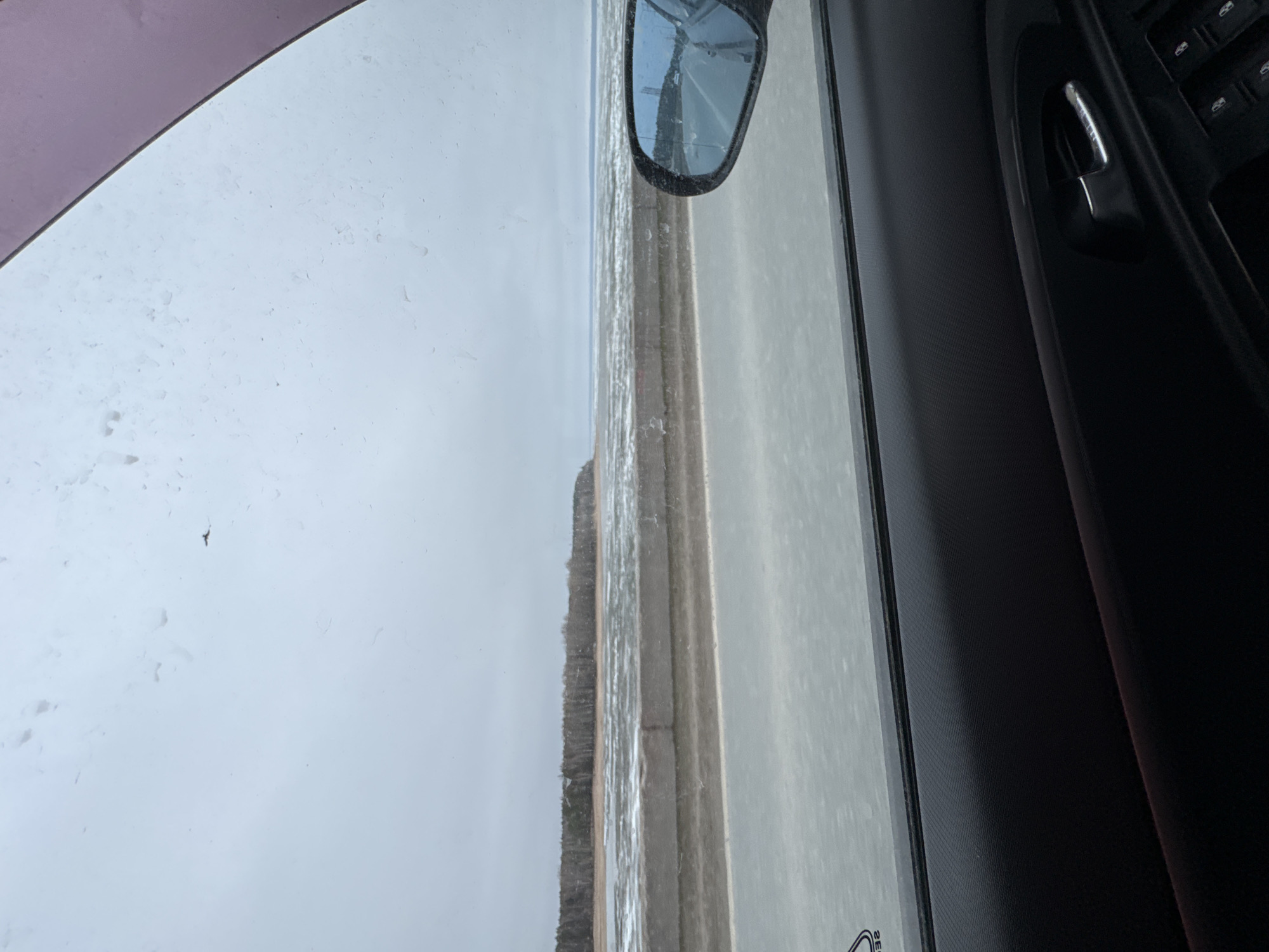 This image, originally rotated 90 degrees clockwise, depicts a desolate, cold landscape viewed from inside a vehicle, likely in Noviy, Russia.
The right side of the frame shows the dark interior of the car's door panel, including a silver door handle, window control buttons, and a side mirror reflecting a partial view of the car's exterior or the blue sky. The car window itself appears speckled with dirt, dust, or moisture.
Looking through the window, a flat, open landscape unfolds under a pale, overcast sky. In the midground, a straight path or road runs parallel to what appears to be a partially frozen body of water, possibly a river or a shore with ice and slush. Beyond this waterway, a distant, dark treeline marks the horizon. The left side of the scene is dominated by a vast expanse of pale sky or snow-covered ground, suggesting winter or early spring conditions. A small, dark speck, possibly a bird or insect, is visible on the window in this area.
No people are visible in the image. The faint reflection of the word "SEE" can be discerned on the window glass near the bottom right. The overall impression is of a quiet, overcast day in a rural or natural setting.
