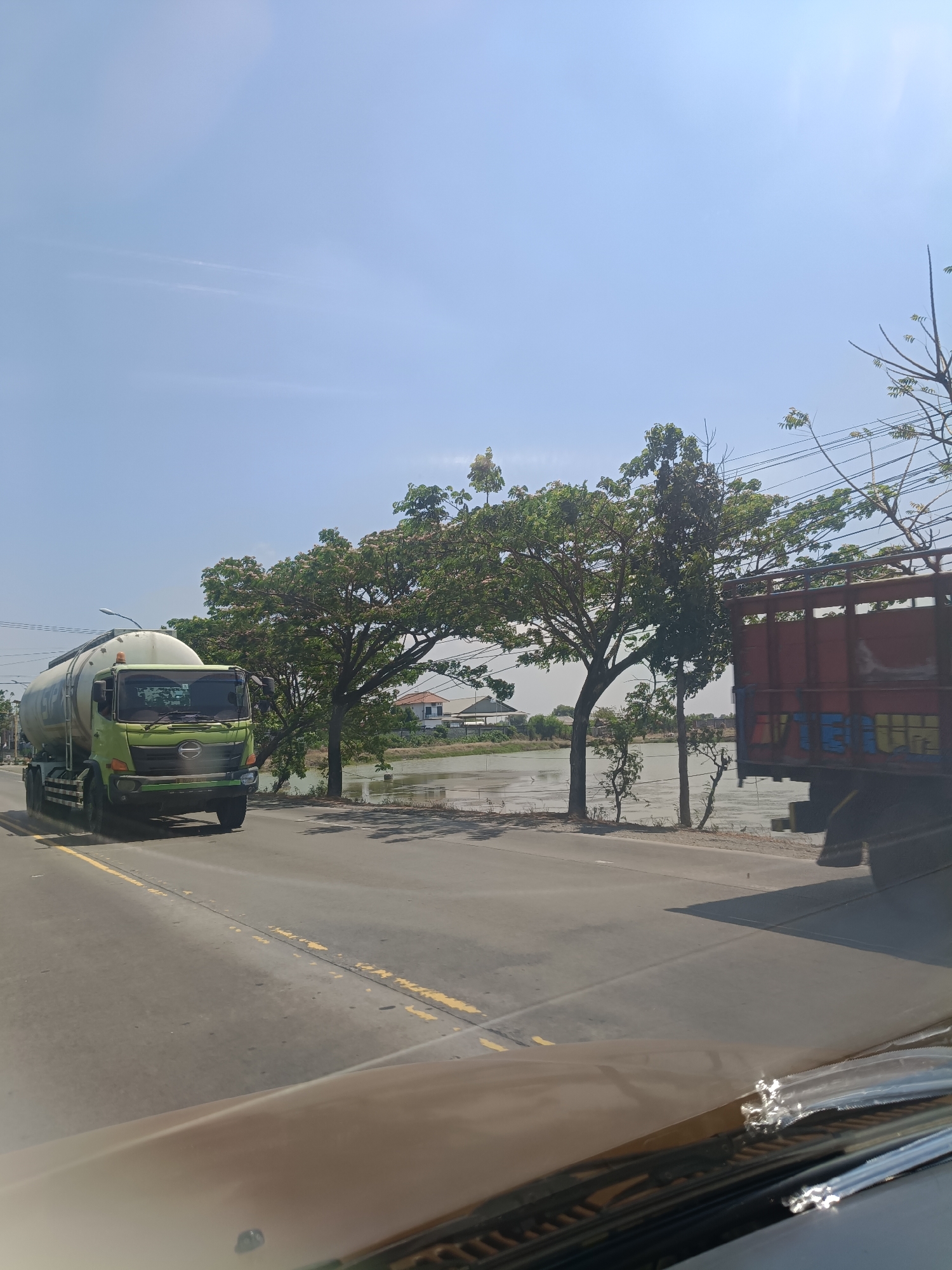 A green tank truck drives on a paved road with yellow lane markings in Duduksampeyan, Indonesia. The truck is on the left side of the road, moving away from the camera. It has a large white tank with some lettering on it. To the right of the road is a body of water with some vegetation and trees along its bank. Further in the background, there are buildings and more trees. Another truck, a flatbed with a red cargo area, is parked or moving slowly on the right side of the road, partially obscuring the view of the background on that side. The sky is clear and blue, indicating a sunny day. The perspective of the image suggests it was taken from another vehicle.