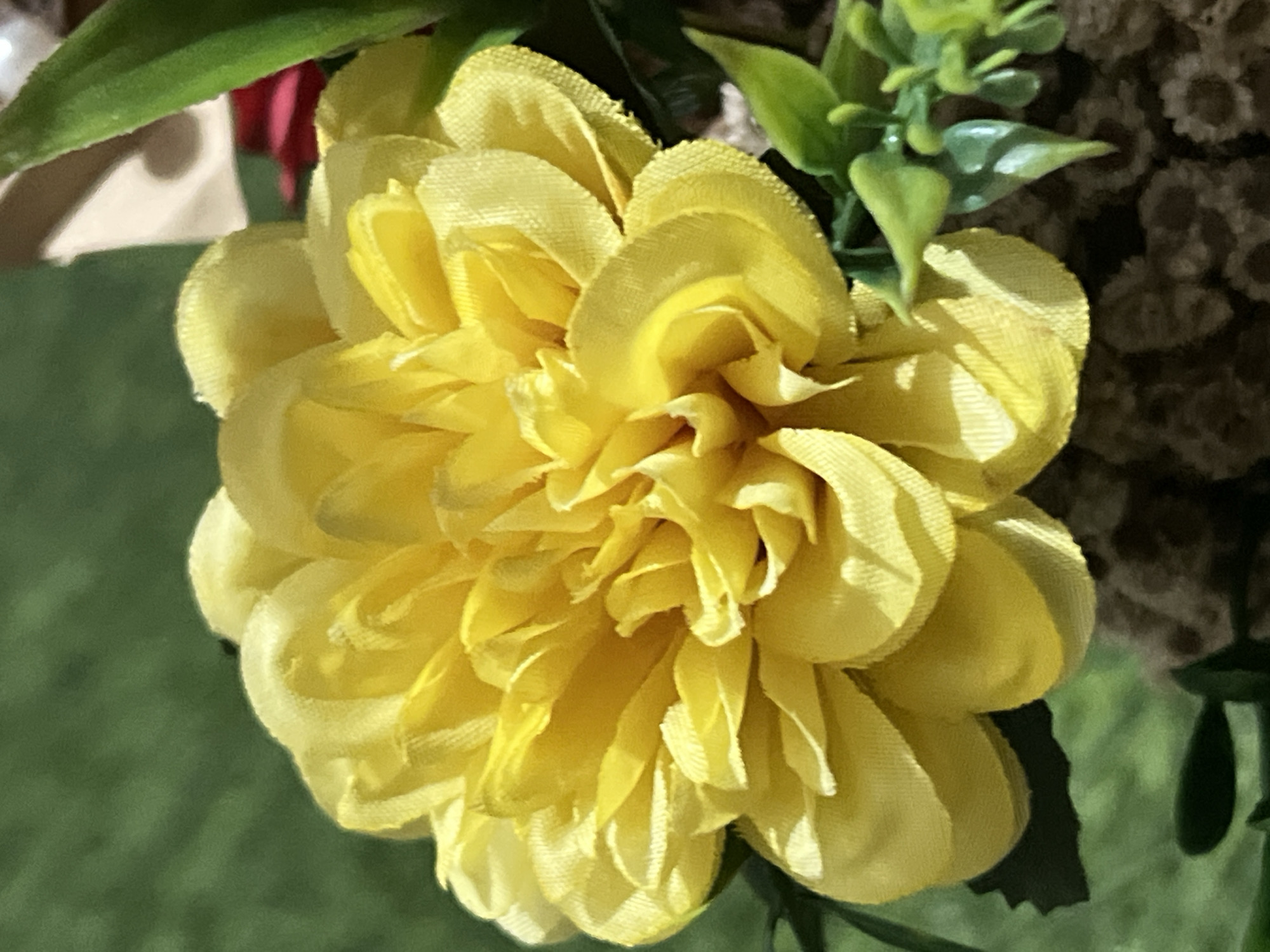 The image is a close-up, overhead shot of a bright yellow artificial flower, likely a chrysanthemum or dahlia, with many layered petals. The petals are textured and have a slightly rough edge. The flower is set against a blurred green background, suggesting foliage or grass. In the bottom right corner, there are hints of other artificial greenery and a dark, textured base or pot. The lighting casts subtle shadows on the flower petals, emphasizing their shape and volume. There is a tiny dark speck, possibly an insect, on the upper left edge of a petal. The scene appears to be a still life arrangement, possibly indoors or in a garden. This image is not specific to Denpasar, Indonesia, and no location in Denpasar can be determined from the visual information provided.