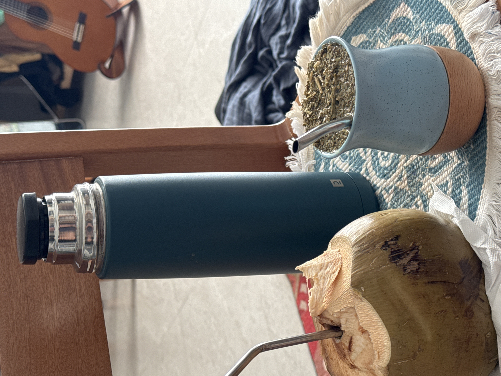 A still life photograph depicts a collection of objects placed on a wooden surface. A blue and brown mate gourd filled with yerba mate and a silver bombilla sits on a patterned woven mat. Next to the gourd, a dark green thermos lies horizontally. A cracked, green coconut with a silver straw inserted completes the arrangement. In the background, a blurry image of a brown ukulele hangs on a wall with a draped dark blue cloth. The location cannot be ascertained based on the image provided.