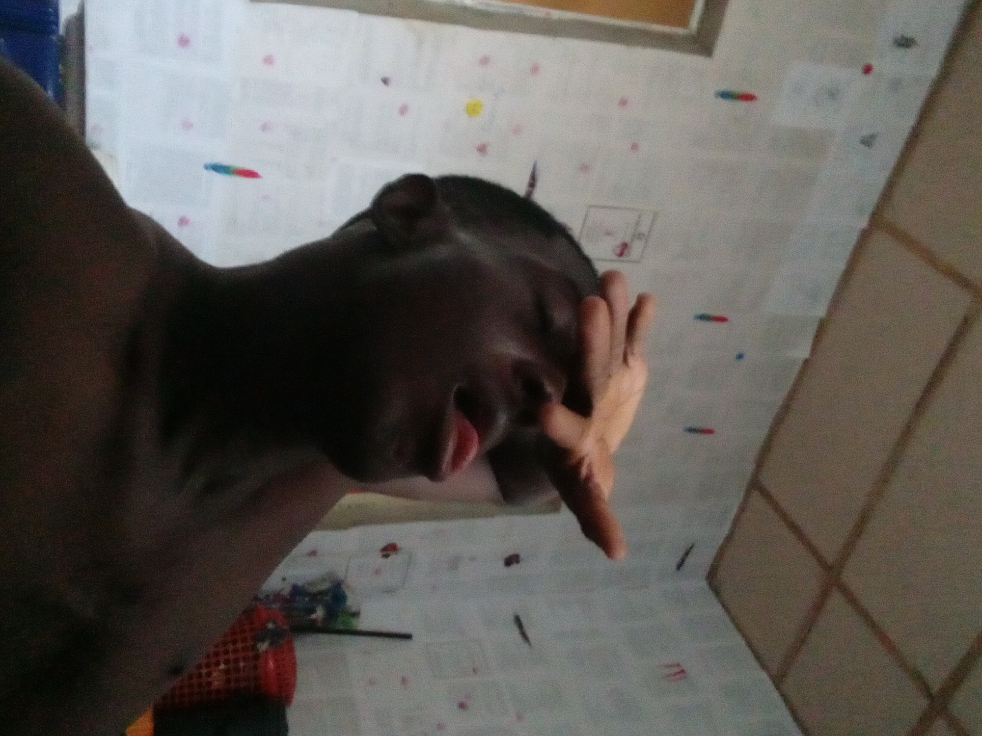 A close-up, tilted shot shows a dark-skinned person with their hand covering the top portion of their face. Part of their cheek, mouth, and chin are visible. Their head is shaved, and their ear is visible. The person's arm extends into the bottom left corner of the image. The background is a white surface with small, colorful markings and what appears to be drawings or notes. A portion of a door or wooden structure with panels is visible on the right side. The scene appears to be indoors. The location of Akwete, Nigeria cannot be verified from the image.