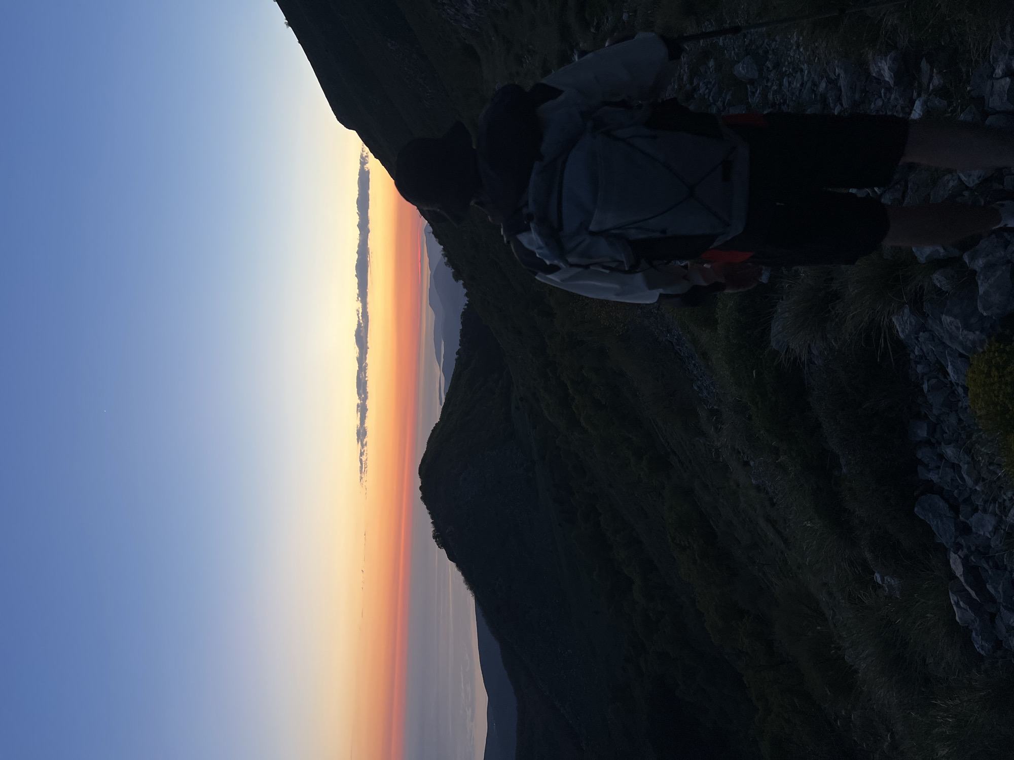 The image depicts a person with a backpack standing on a rocky mountain slope in Dolovi, Montenegro, likely observing a sunset. The sky transitions from blue to orange near the horizon, marked by a narrow cloud formation. In the distance, shadowy mountain ranges are visible. The foreground features dark vegetation and rocks, suggesting a mountainous environment. The individual appears to be stationary, possibly taking in the view.