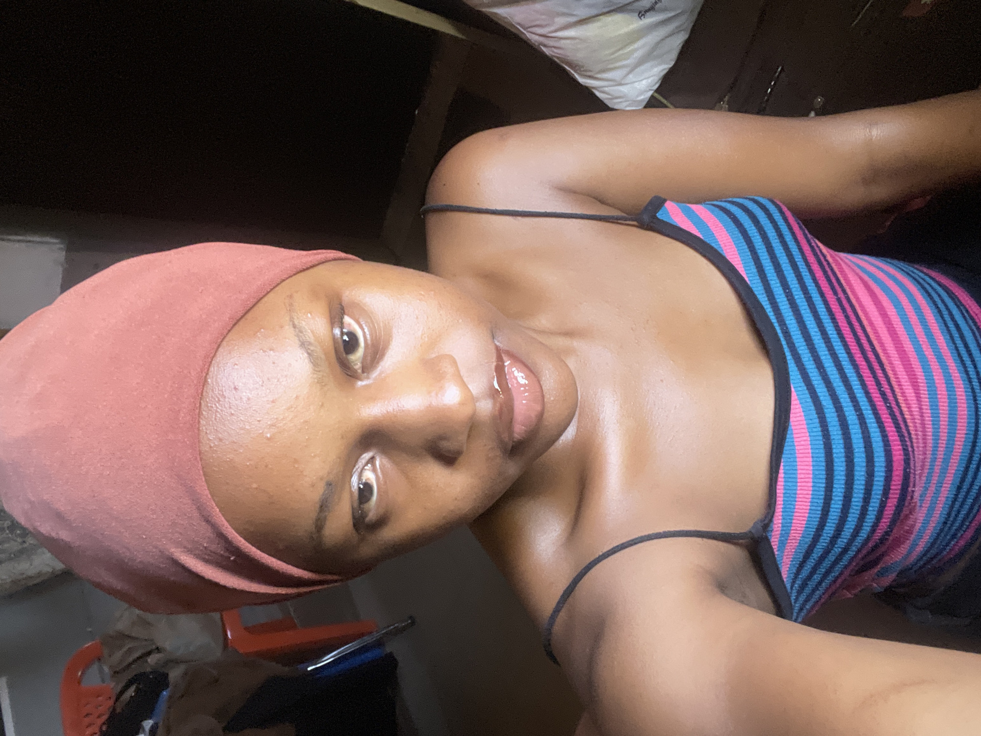 The media file is a close-up, vertically oriented selfie of a young woman. She is wearing a pink headscarf and a striped tank top with blue and pink lines. Her skin appears dewy, suggesting it may be humid, consistent with the tropical climate of Jos, Nigeria. Her lips are glossy, and she has a relaxed expression, possibly indicating a casual or private moment.
In the background, there are hints of an indoor setting. A white, bagged object is visible in the upper right corner. Other objects, including what appears to be furniture, are blurred and out of focus. The lighting is dim, suggesting it might be evening or nighttime. There is no discernible weather information.
The subject appears to be posing for the camera, with her gaze directed towards it. The overall mood is intimate and personal. No visible text is present in the image.