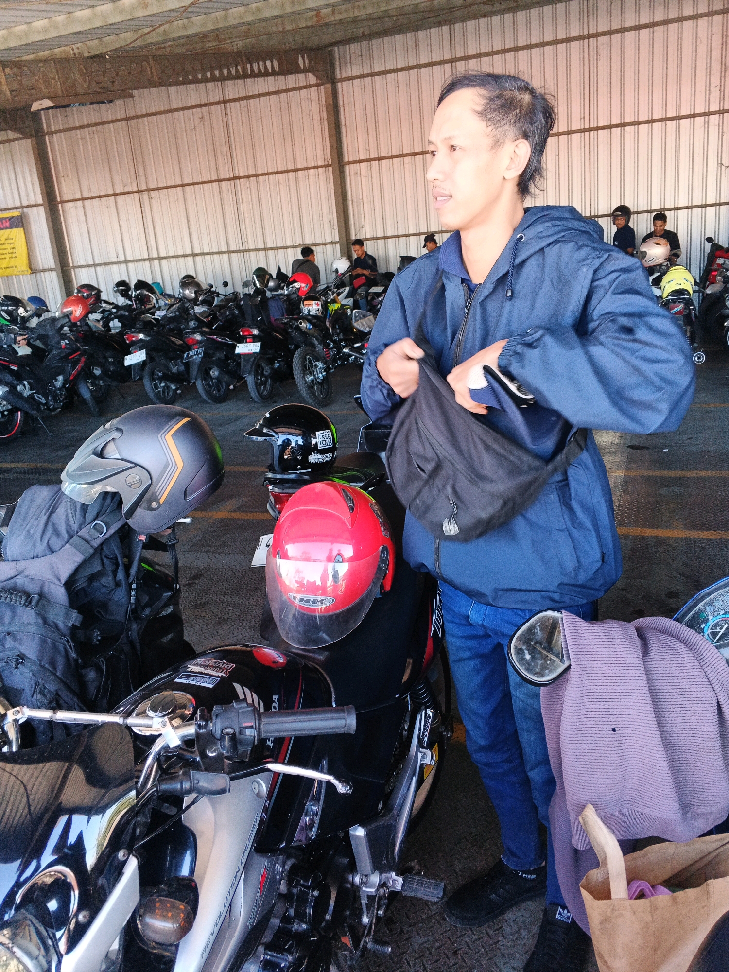 A man in a blue jacket and jeans stands in a covered parking area, likely a motorcycle parking lot, given the numerous motorcycles parked behind him. He is reaching into a black sling bag across his chest. In the foreground, three motorcycle helmets are visible: one grey, one black, and one red. A black backpack is placed on a motorcycle seat. The background shows rows of parked motorcycles and a corrugated metal wall. The overall setting suggests an outdoor urban environment in Banjararum, Indonesia, possibly a transportation hub or a public facility.