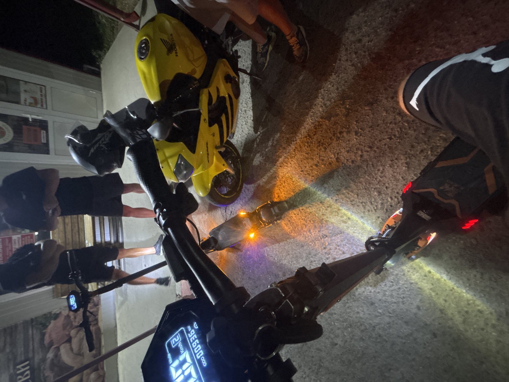In Marten, Bulgaria, on a paved surface at night, two electric scooters and a yellow Honda CBR motorcycle are parked. The perspective is from the rider of the foremost scooter, which has a digital display showing "Mode" and "Kmh" with a reading of "00936 km". The second scooter, behind and to the left, has its brake lights illuminated. A yellow Honda CBR motorcycle, with the "HONDA" logo visible, is to the right. Several individuals, whose legs and lower torsos are visible, are standing near the motorcycle. One person is wearing black shorts with white stripes. The background includes a building with glass doors and a large poster.