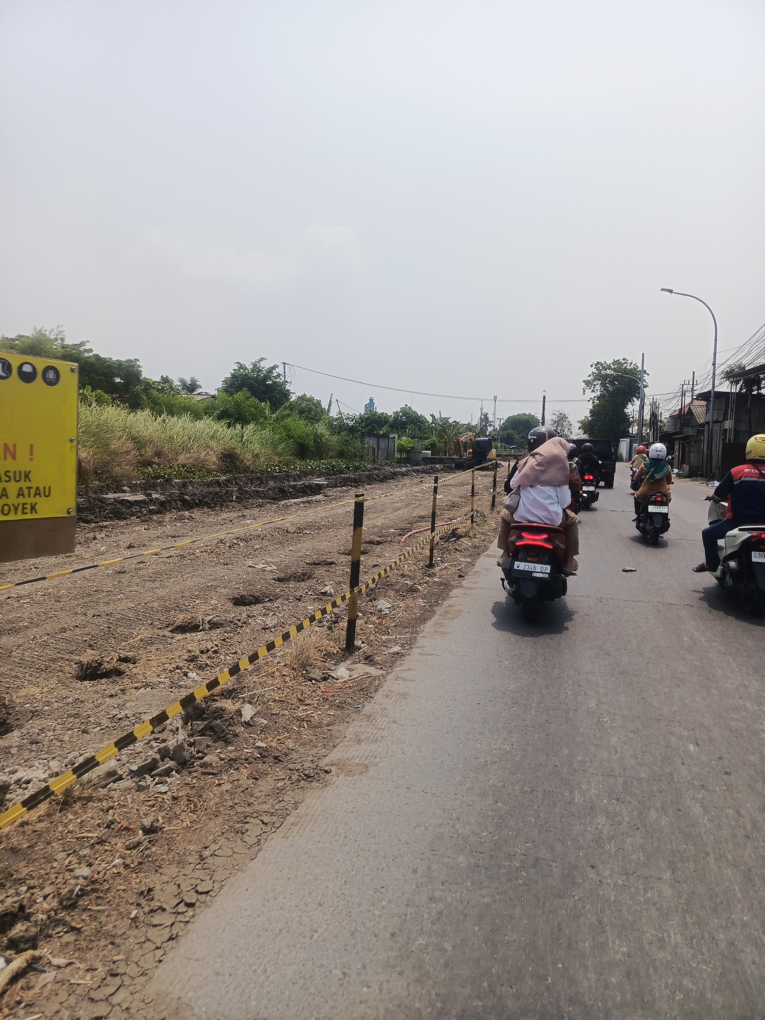 In Surabaya, Indonesia, an outdoor scene depicts a construction zone alongside a paved road. The foreground shows a dirt path with scattered debris and a yellow and black striped caution tape barrier. Several vehicles, including motorbikes carrying passengers, are traveling on the road. On the left side of the road, a yellow sign with black text is visible, partially obscured by foliage and a construction barrier. In the background, there is dense greenery, overhead power lines, and residential buildings. The sky is overcast and hazy.
