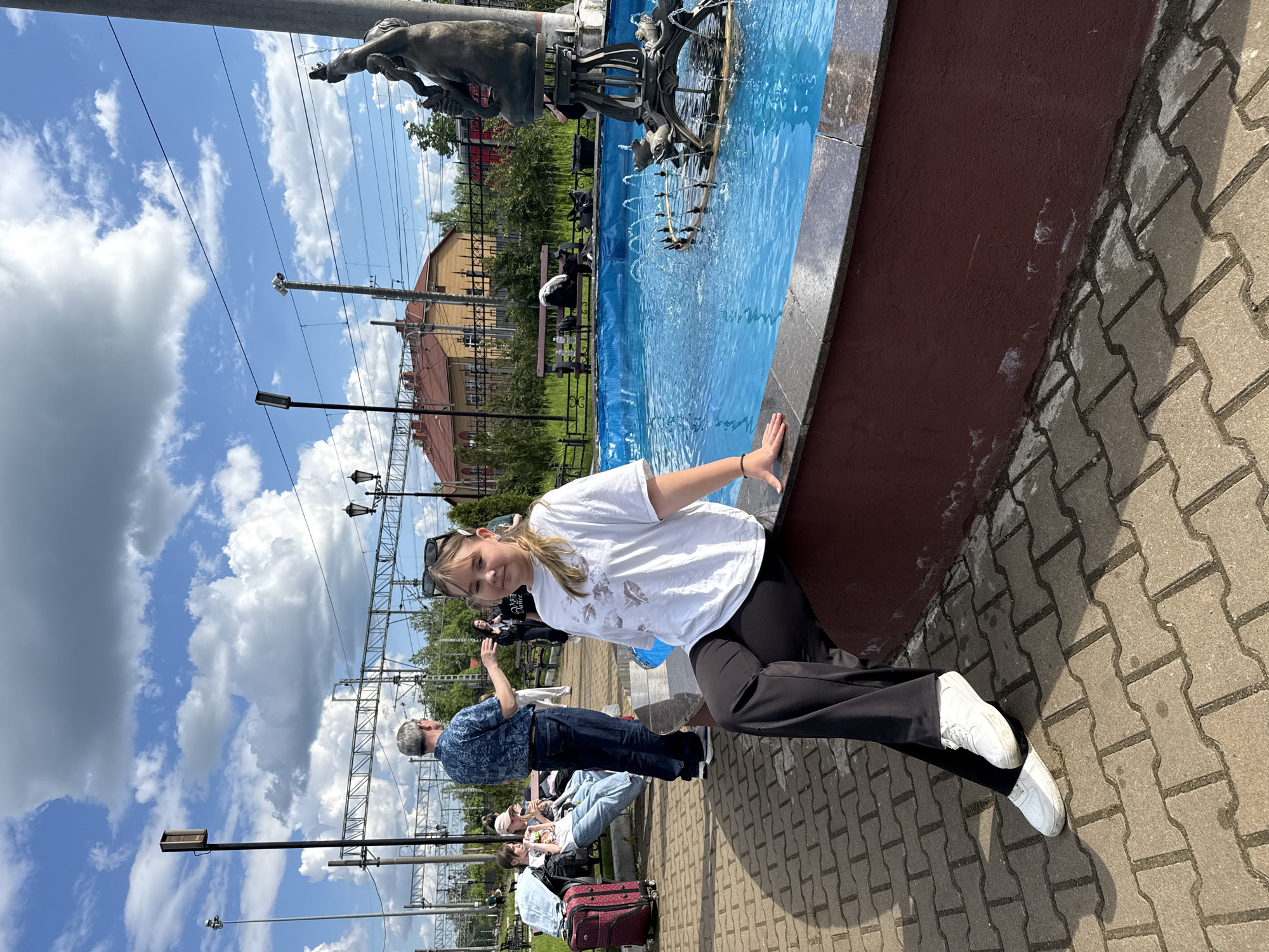 The image, correctly oriented, depicts a bright, sunny outdoor scene in Malaya Vishera, Russia, likely near a train station. A young woman with blonde hair, wearing a white t-shirt, black trousers, and white sneakers, is the central subject. She smiles faintly at the camera, leaning on the dark red edge of a large, ornate fountain. Sunglasses rest on her head.
The fountain features vibrant blue water and intricate dark metal sculptures depicting human-like figures and possibly marine life, with water jets creating splashes. In the background, several other people are visible; one man in a blue patterned shirt stands with his back to the camera, gesturing, while others are seated on benches, some next to a red suitcase.
The ground is paved with interlocking grey concrete pavers. Above, a clear blue sky with scattered white, fluffy clouds indicates bright, mid-day sunlight. Numerous railway overhead power lines and support structures, along with streetlights, span the scene, confirming the proximity to a railway line. A building with an orange-red roof and a decorative black fence are also visible in the distance. No legible text is discernable.