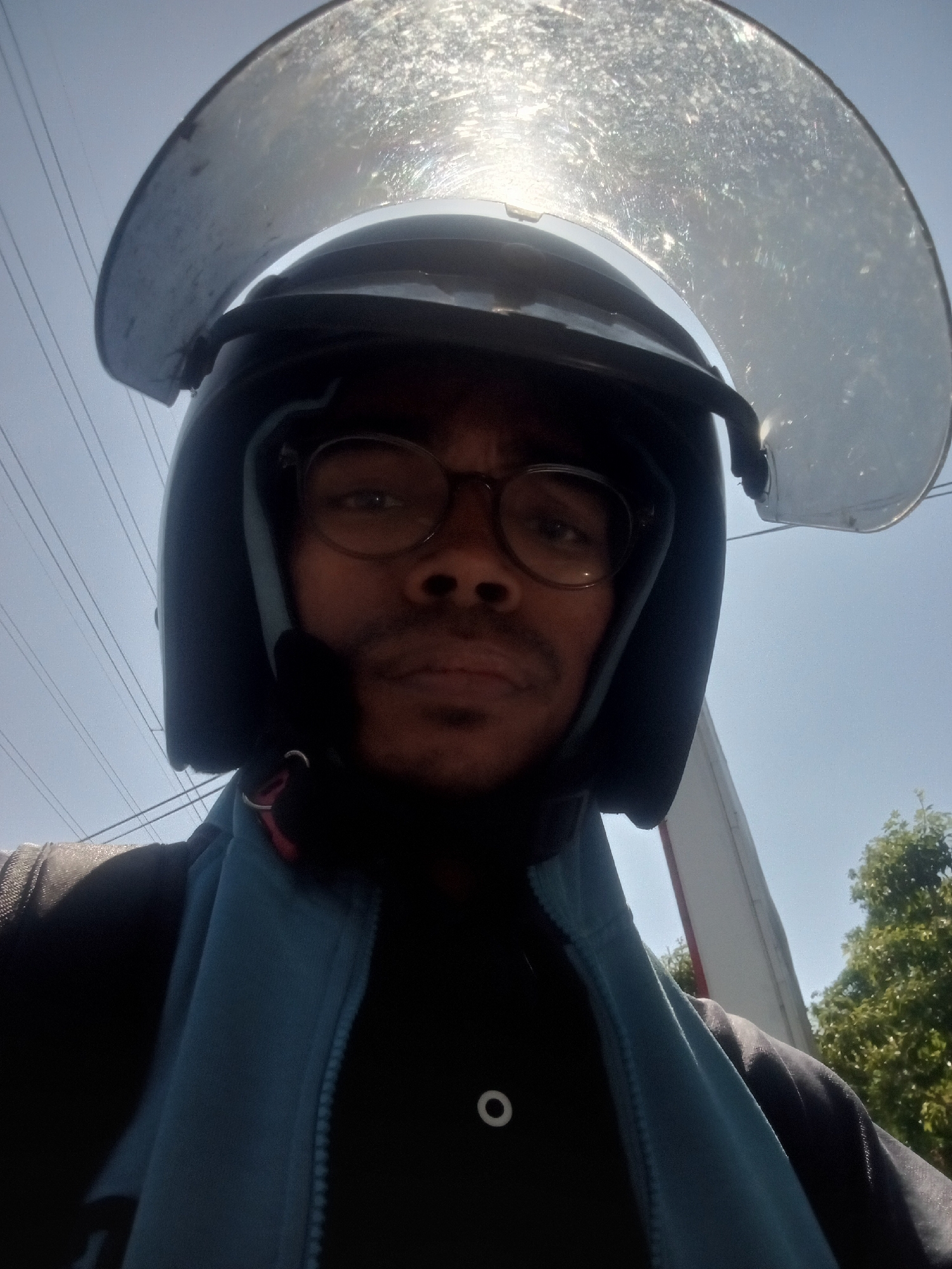 A close-up, upward-facing selfie of a young man wearing a black helmet with a clear visor that is partially obscured by sunlight. He is also wearing round glasses and a light blue zippered jacket over a dark shirt. The man appears to be outdoors, with a clear blue sky and power lines visible in the background. Trees and a building are also present. The image is shot in a selfie style, with the man's face taking up most of the frame. No specific location in Surabaya, Indonesia, can be identified from the visual information provided.