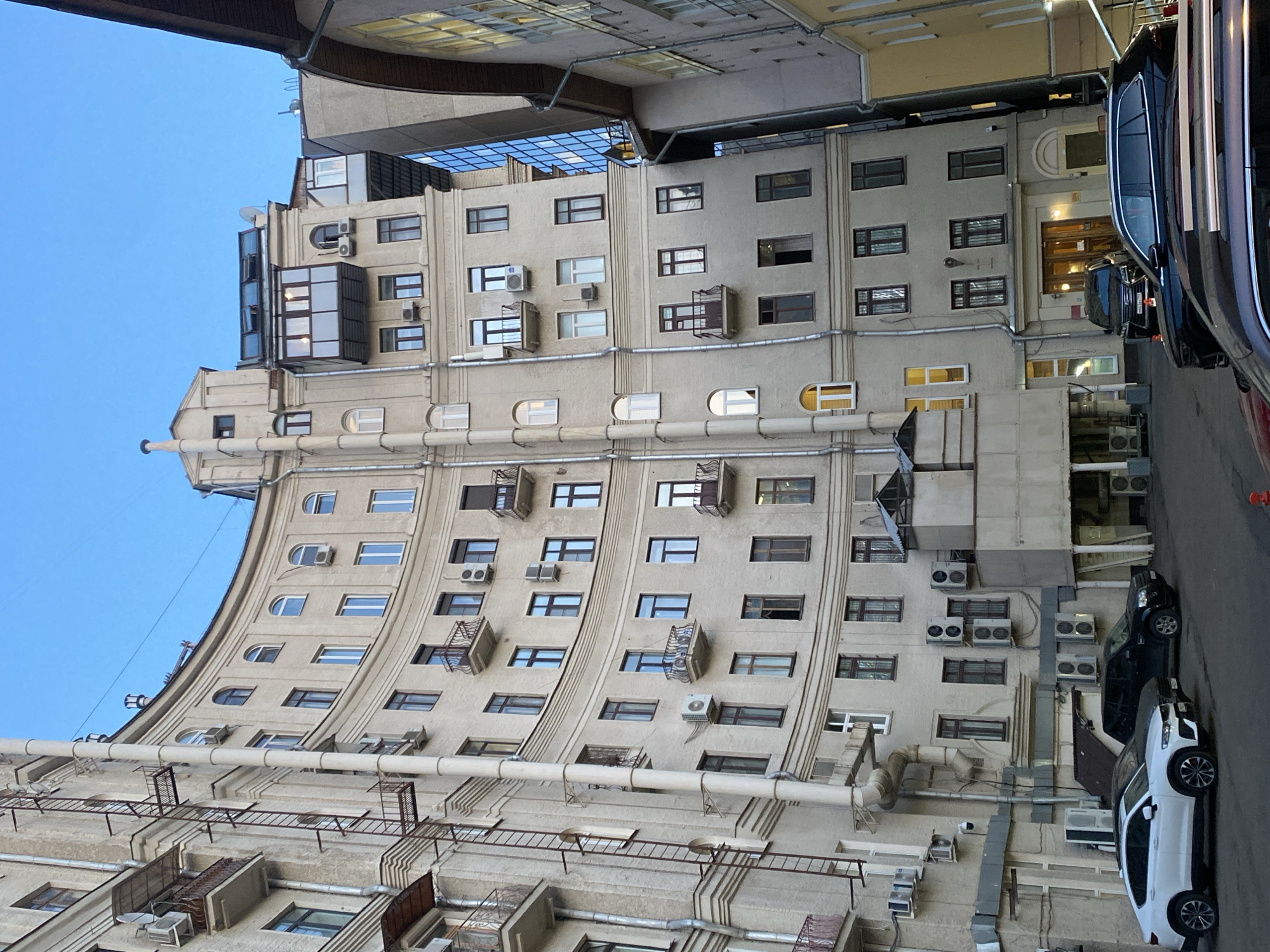 The image captures a multi-story building in Moscow, Russia. The building has a curved facade with numerous windows and air conditioning units attached to its exterior. A large pipe runs along the building's lower section, supported by metal frames. Adjacent to the building is a narrow street with parked cars. The sky is clear blue. The angle of the photograph suggests it was taken from a low vantage point, looking upwards.