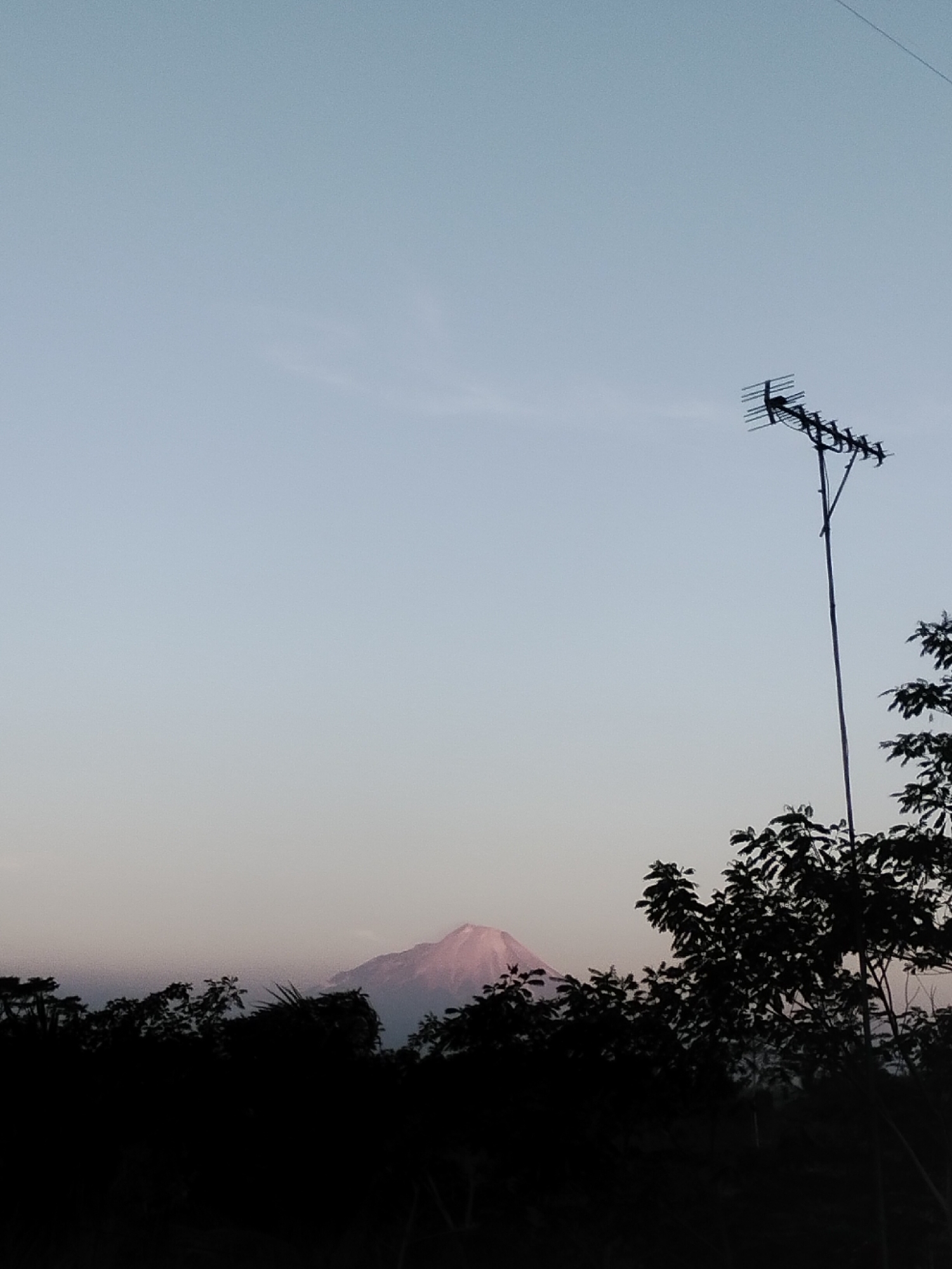 The image depicts a serene landscape in Magelang, Indonesia, at what appears to be either dawn or dusk, given the soft, diffused light. In the foreground, a dense silhouette of dark foliage, likely trees and bushes, spans the lower portion of the frame, obscuring the ground and any potential human activity. Dominating the right side of the image is a tall, slender pole supporting a prominent television antenna, its metallic structure silhouetted against the sky. In the mid-ground, a mountain, likely Mount Merapi due to its distinctive conical shape and location relative to Magelang, is visible. Its peak is softly illuminated, suggesting the low angle of the sun. The sky above is a gradient of pale blue, transitioning to a lighter, almost white hue near the horizon, indicating the absence of harsh sunlight. The overall atmosphere is one of tranquility and natural beauty, with man-made elements like the antenna providing a contrasting visual feature to the natural landscape. There are no people or animals visible in the image.
