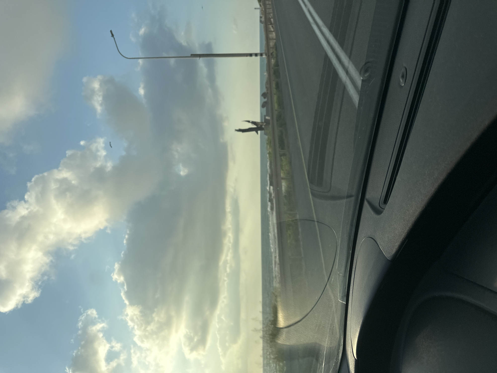 The image captures a view from inside a vehicle, looking out through the windshield. The scene depicts a coastal road in Nahariyya, Israel. The road is paved with white lines marking the lanes. To the left of the road, a body of water, likely the Mediterranean Sea, is visible, with gentle waves breaking near the shore. A metal guardrail separates the road from the sea. In the distance, along the coastline, are some sparse green vegetation and a tall streetlamp. Above, the sky is partly cloudy with large, white clouds illuminated by the sun. A dark, abstract sculpture, possibly a bird-like form, stands near the roadside. The windshield has some smudges and reflections, indicating it is daytime and likely sunny. The dashboard of the vehicle is visible in the foreground, partially obscuring the view.