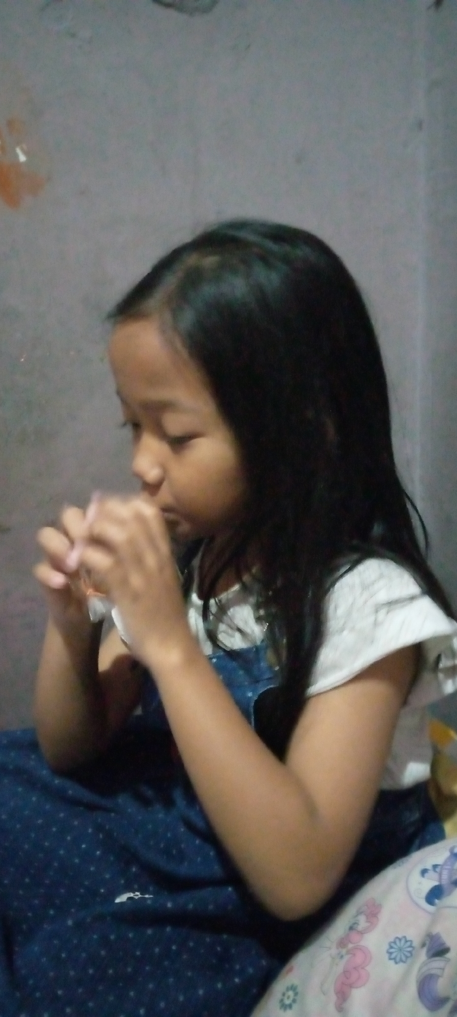 A young girl, with dark hair and eyes, is seated indoors, facing left. She is wearing a white ruffled shirt and a dark blue dress with white polka dots. Her hands are held up to her face, and she appears to be holding a small, light-colored object. The background consists of a plain, textured wall and a patterned pillow. The lighting is dim. The image is a medium shot.