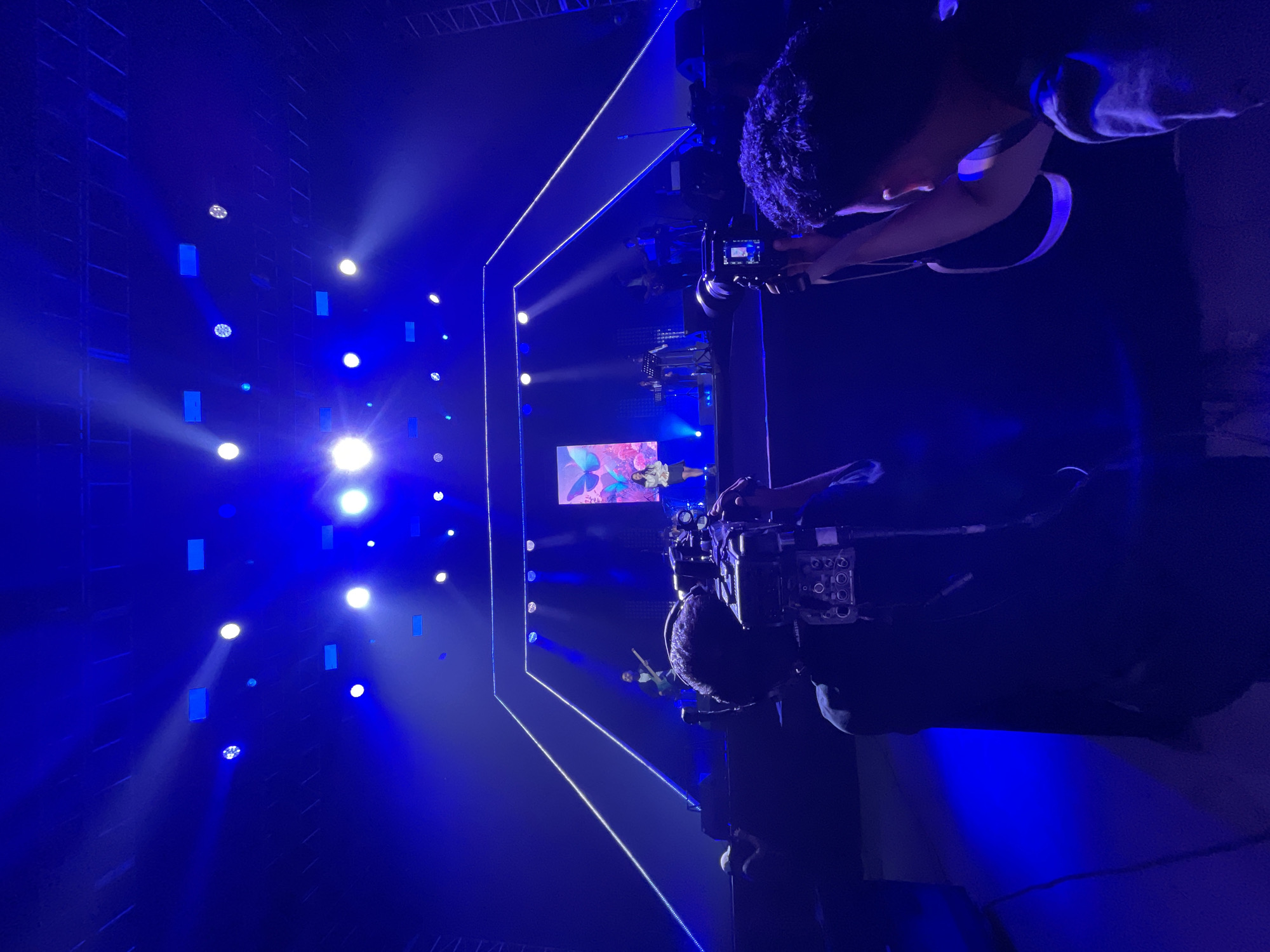 The image depicts a concert stage in Salembaran, Indonesia. The stage is illuminated with blue lights and features a large screen displaying a performer. Several cameramen are positioned to capture the event, focusing their lenses on the stage. The environment is dark except for the stage lighting, highlighting the performers and the technology used to film them.