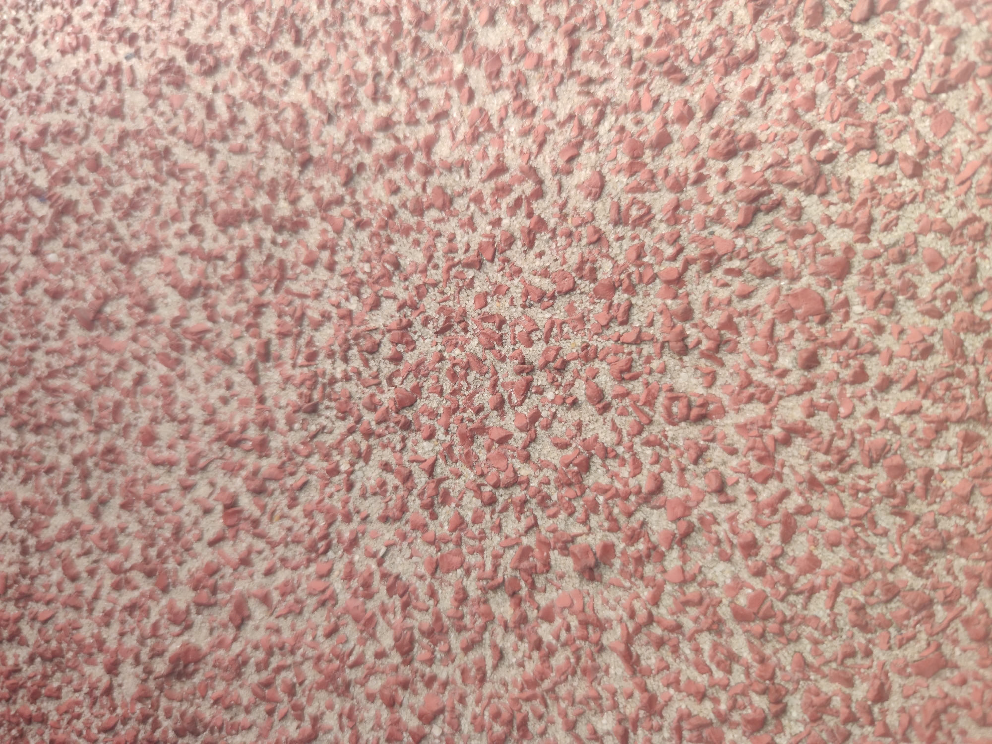 The image shows a close-up, abstract view of a textured surface. It appears to be an outdoor surface, possibly a walking path or playground, composed of small, irregularly shaped reddish-pink granules embedded in a lighter, sandy-colored matrix. The granules are varied in size and shape, creating a rough, uneven texture. The lighting suggests it is daytime, with shadows subtly defining the texture. There are no people or identifiable objects within the frame. The image is likely a detail shot, focusing solely on the material composition and texture. The location in Surabaya, Indonesia, is not visually discernible from the provided image alone.