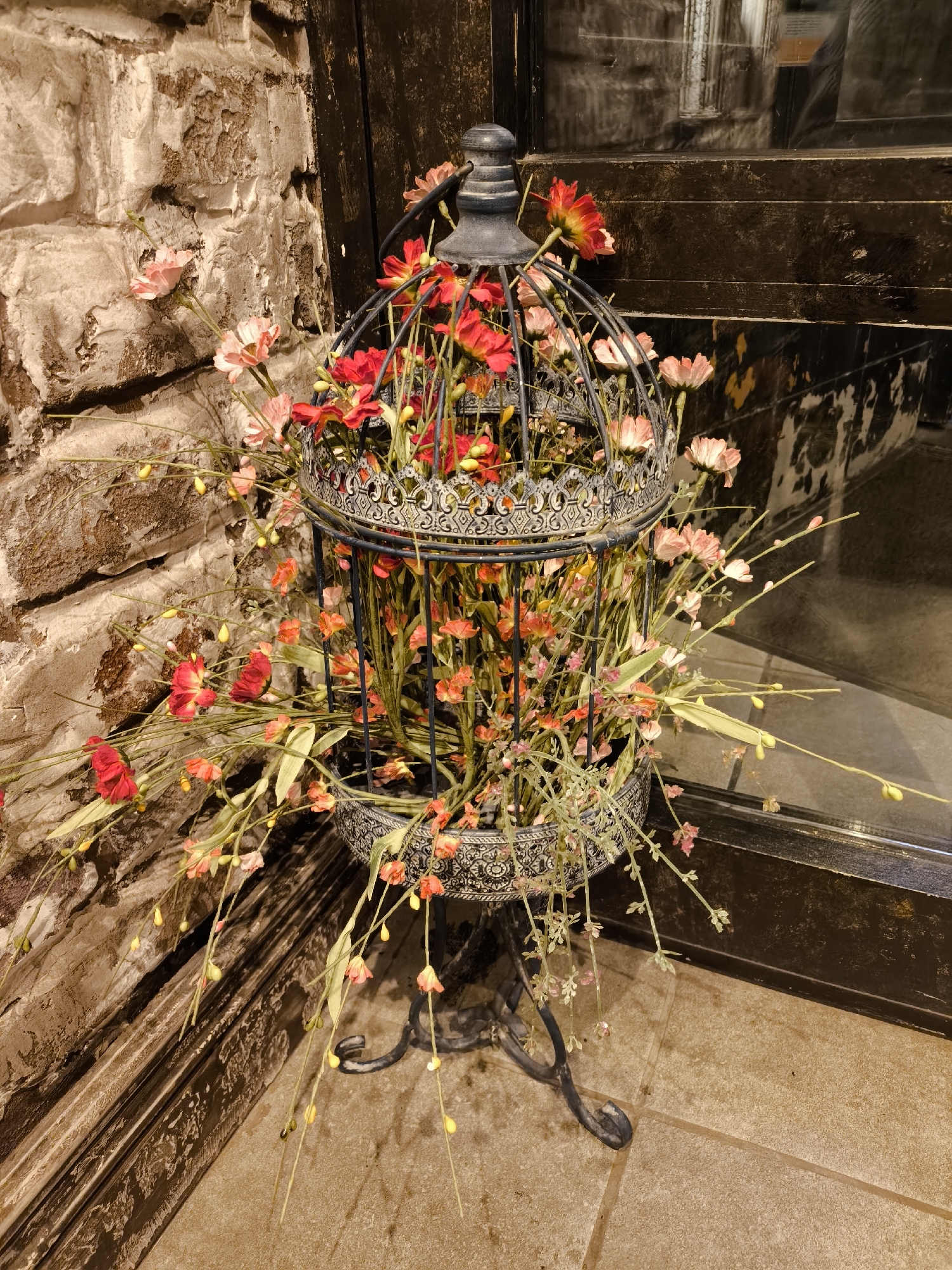 A decorative birdcage stands on a tiled floor adjacent to a brick wall and a dark, reflective door or window. The birdcage is filled with faux flowers in shades of red, pink, and orange, with some extending beyond the cage's structure. The birdcage itself is mounted on a wrought iron stand with curved legs. The environment suggests an interior space, possibly a shop or restaurant. The location is not identifiable as Moscow, Russia from the visual information provided.